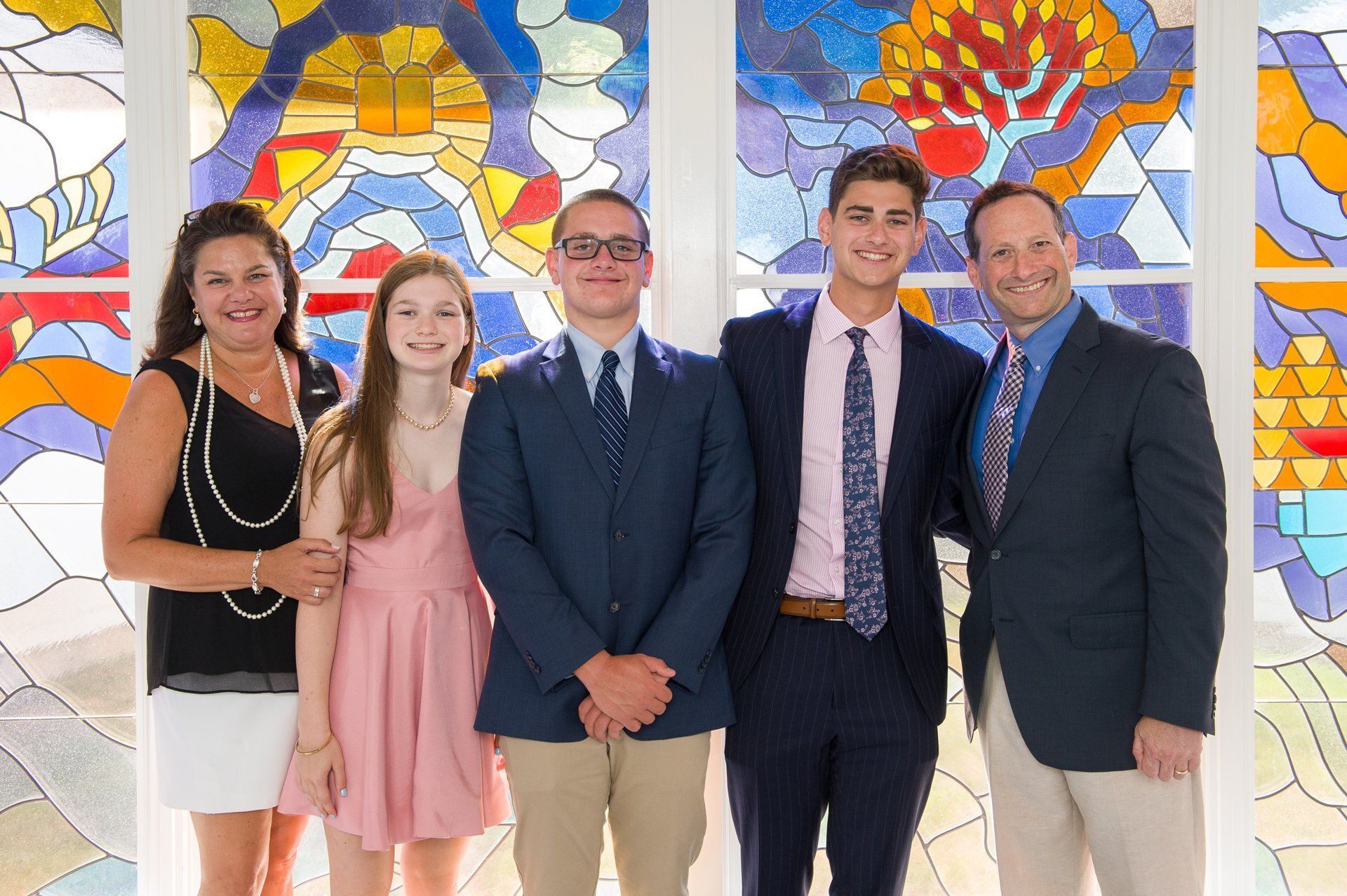 Five people posing indoors in front of colorful stained-glass artwork.
