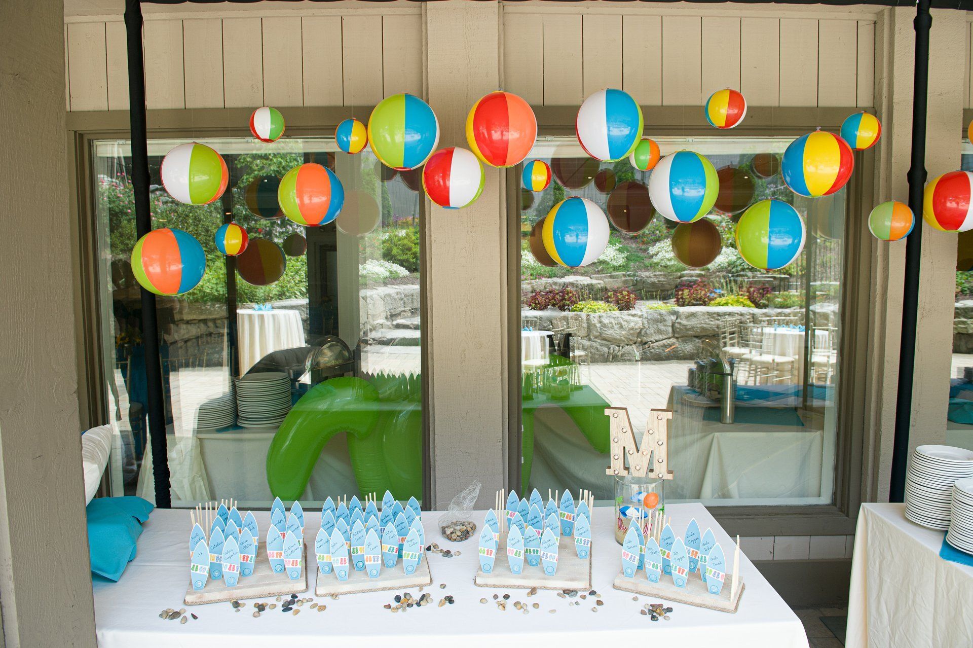 Outdoor party table with colorful balloons, blue water bottles, and a large silver “M” decoration
