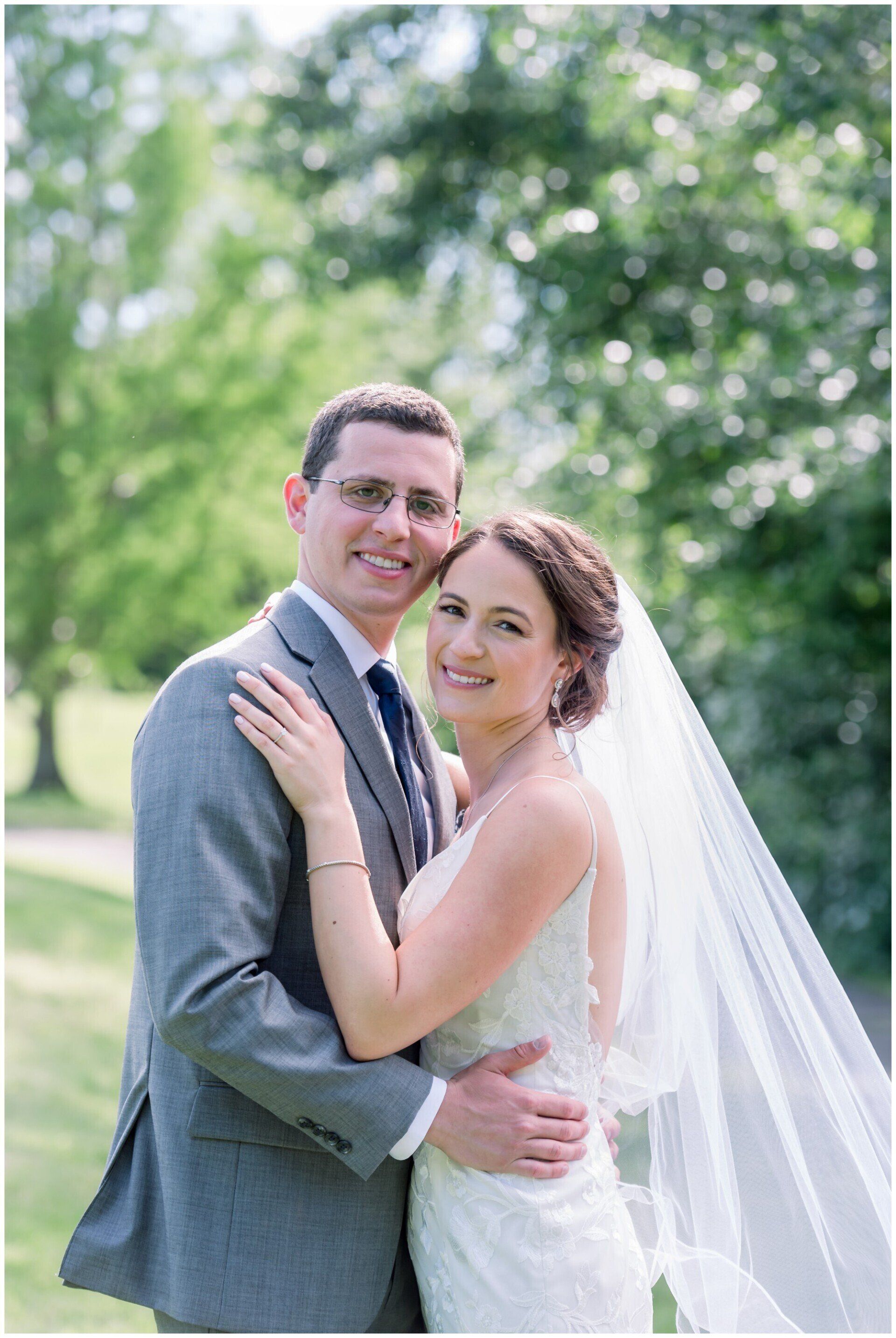 Bride and groom embracing outdoors, smiling in wedding attire with a veil flowing behind them