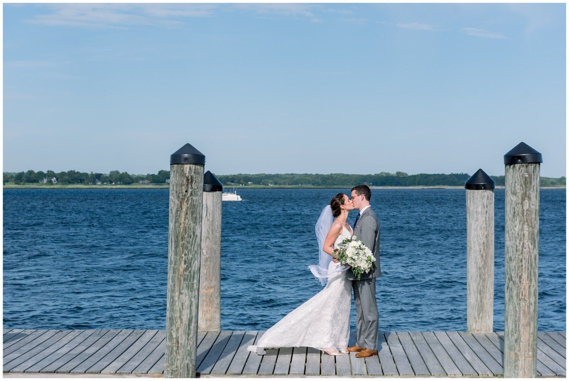 Bride and groom kiss on a dock by the water, with blue sky and wooden posts.