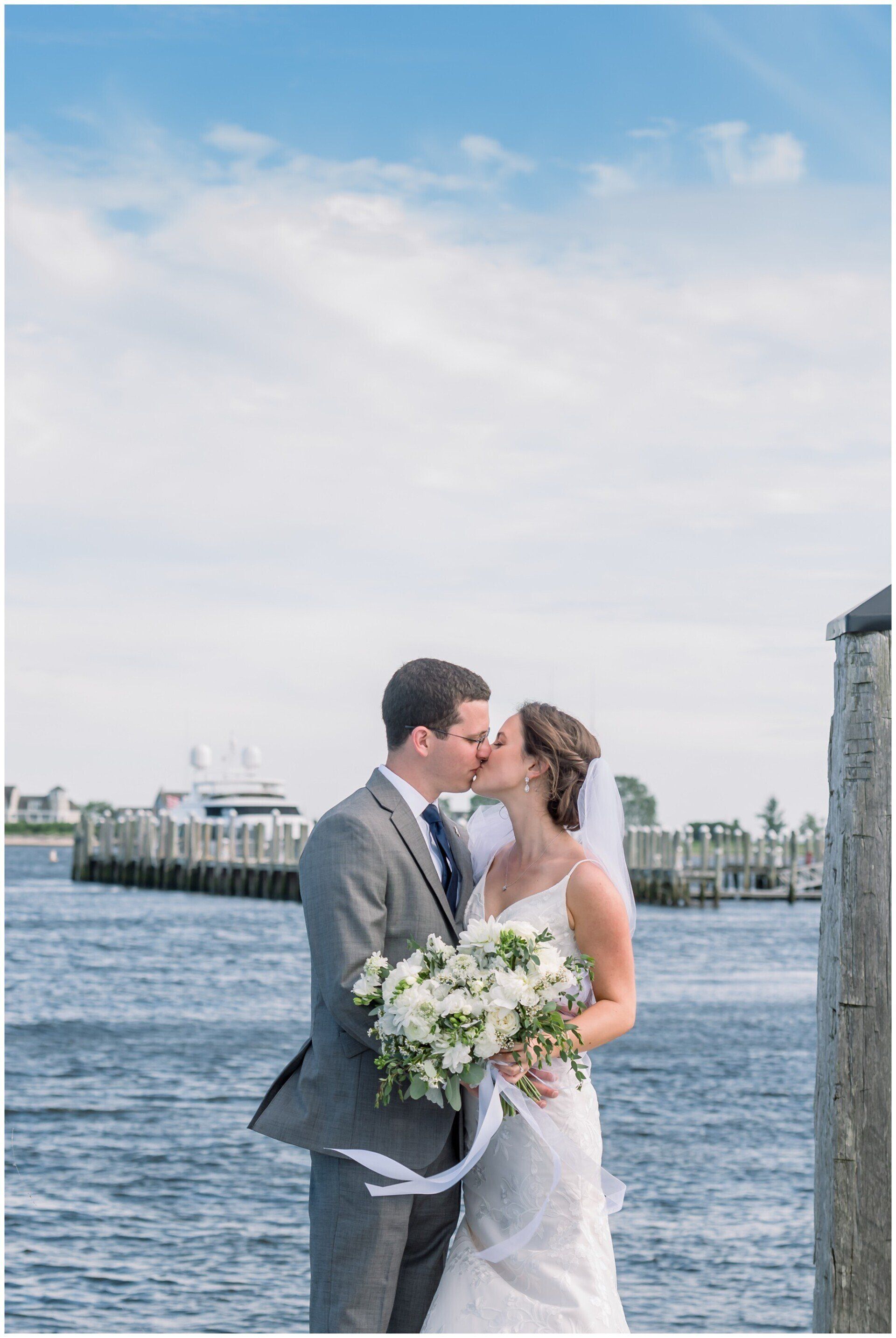 Bride and groom kiss by the waterfront, holding a bouquet under a bright sky
