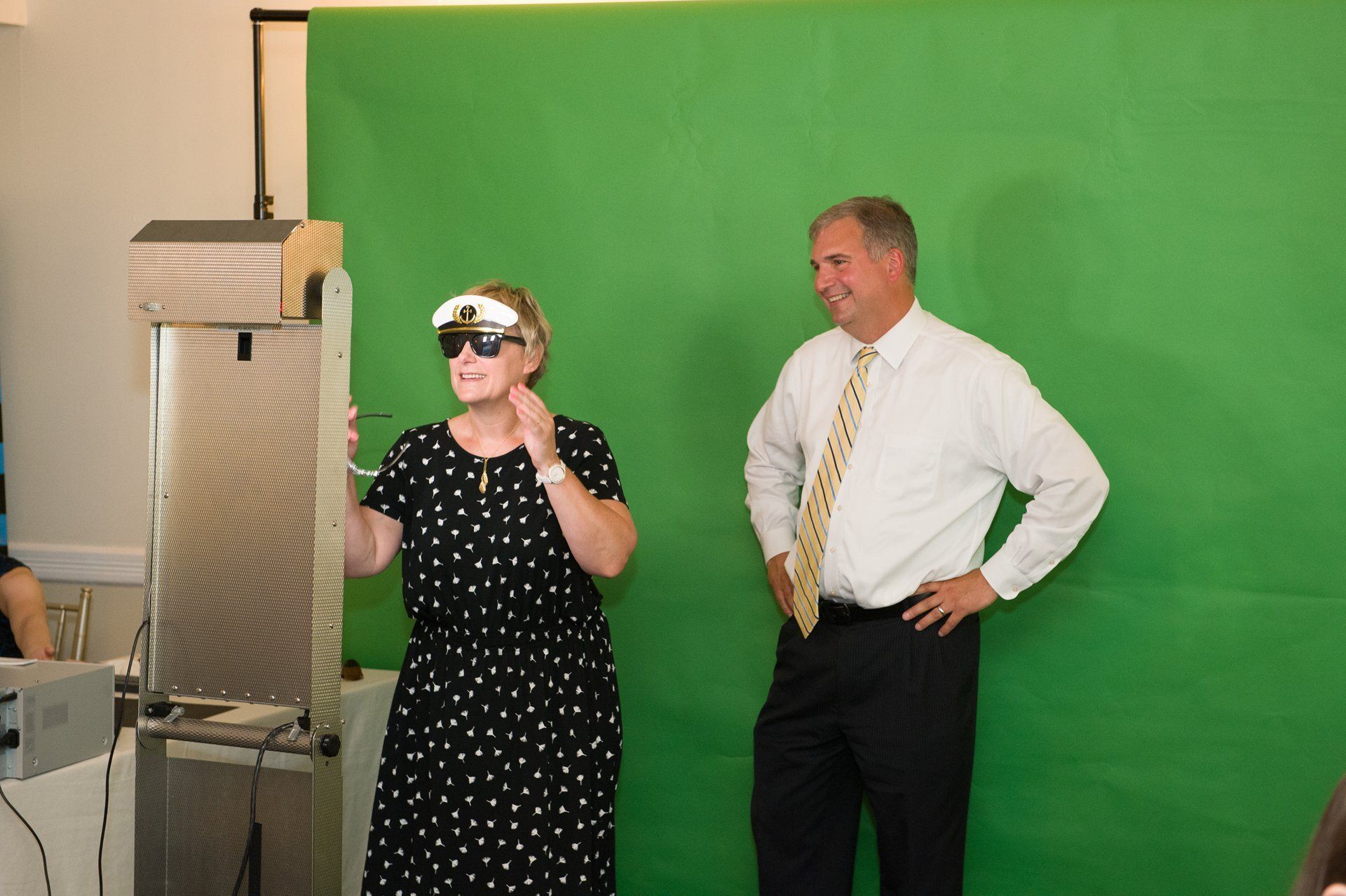 Two people standing before a green screen, one wearing VR goggles and gesturing, the other posing with hands on hips.