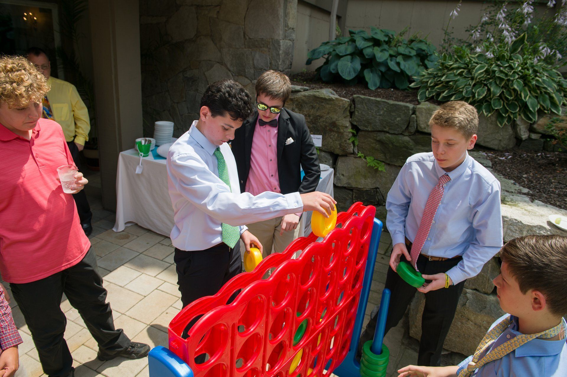 Children standing around a red Connect Four game outdoors, taking turns dropping colored disks