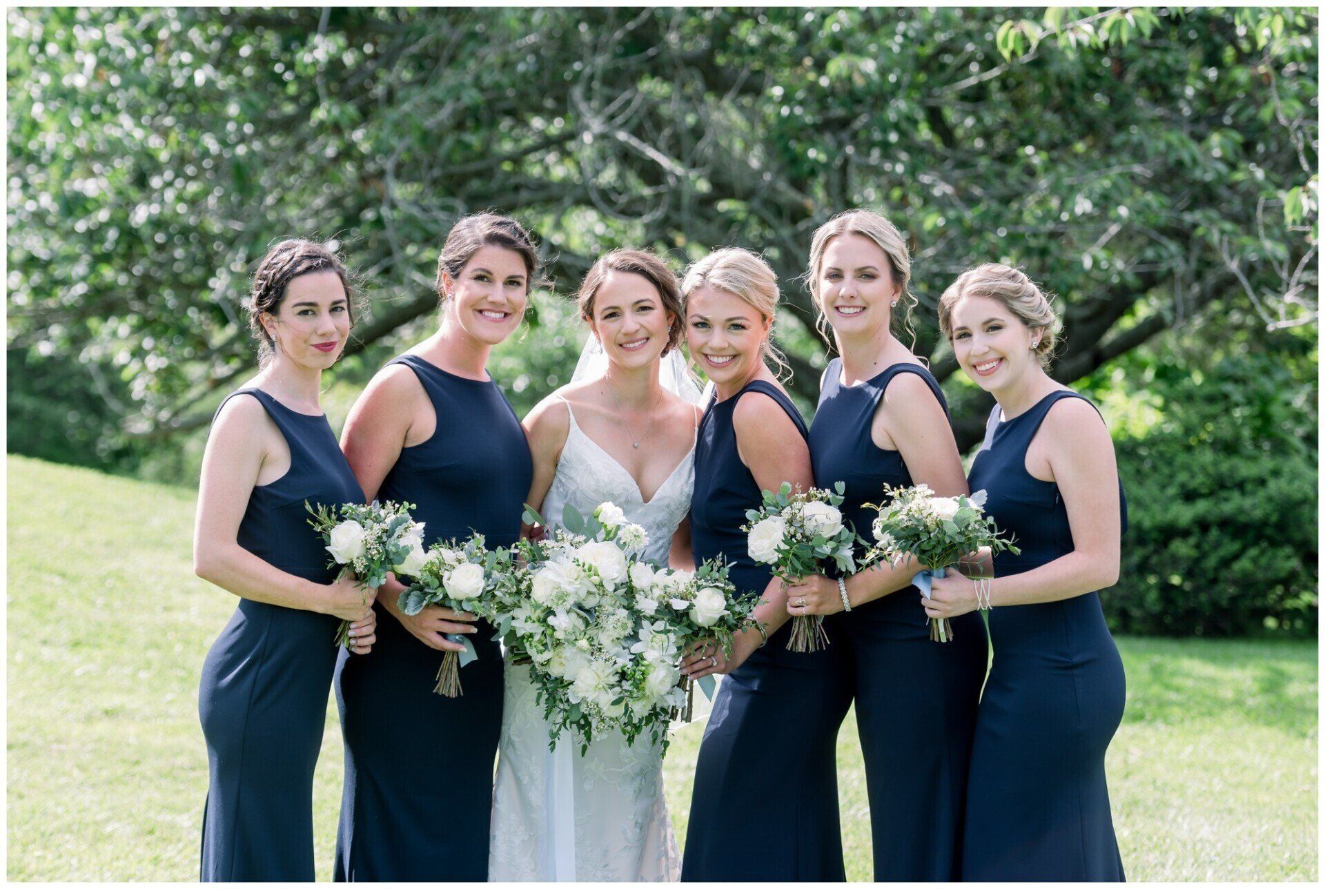 Six women in navy dresses and one in a white dress stand outdoors holding bouquets.