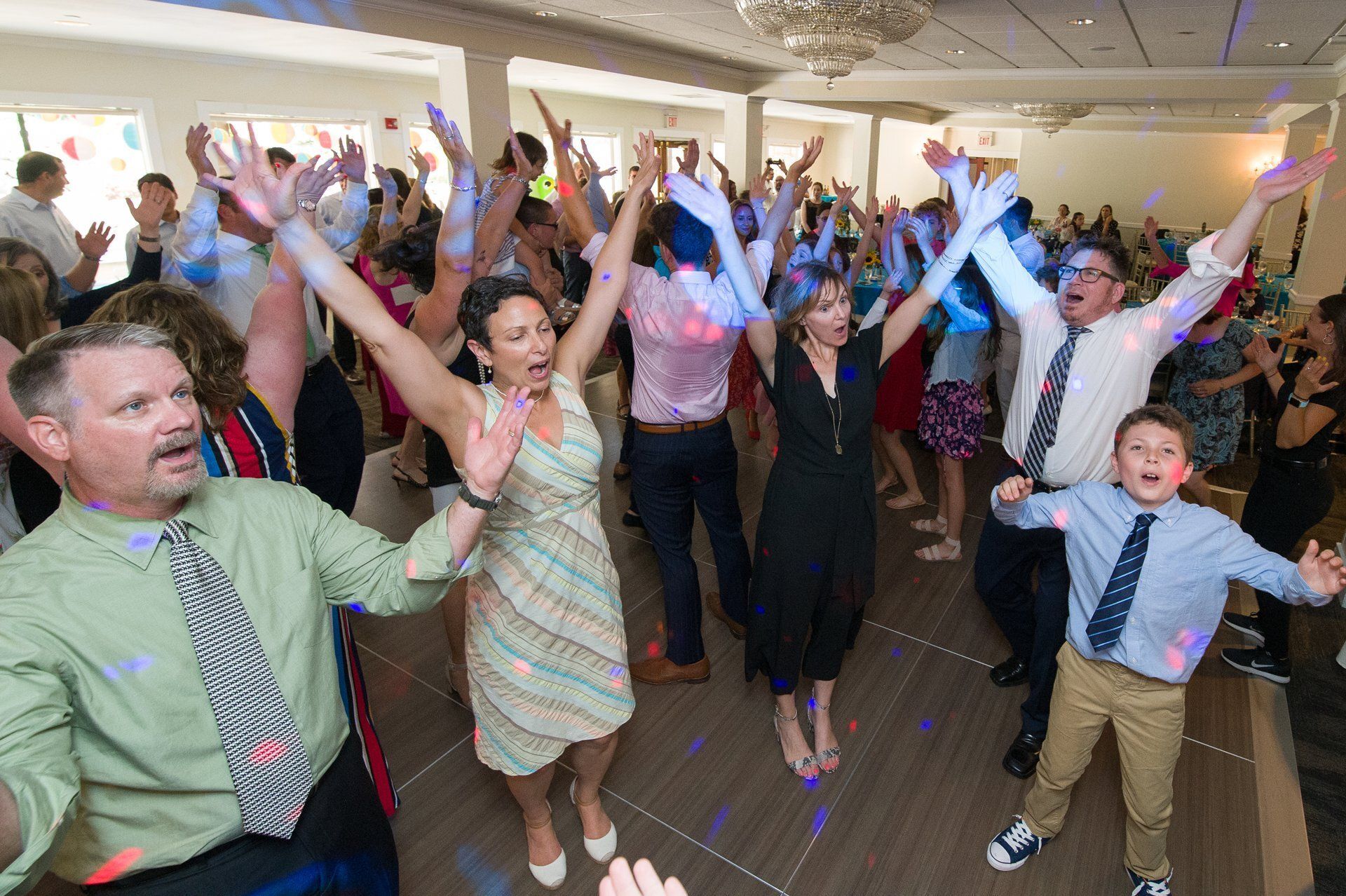 People dancing with arms raised at a lively indoor party, smiling in a banquet hall.