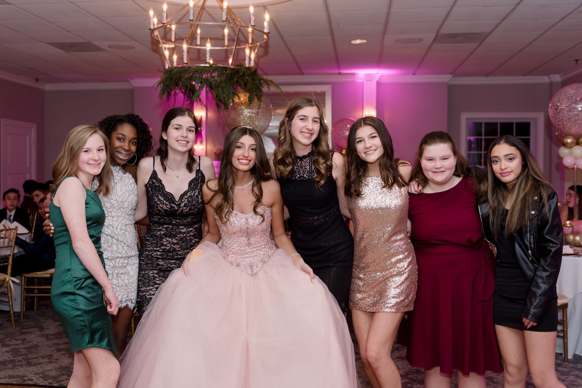 Seven people posing in formal dresses at a pink-lit celebration venue, smiling together.