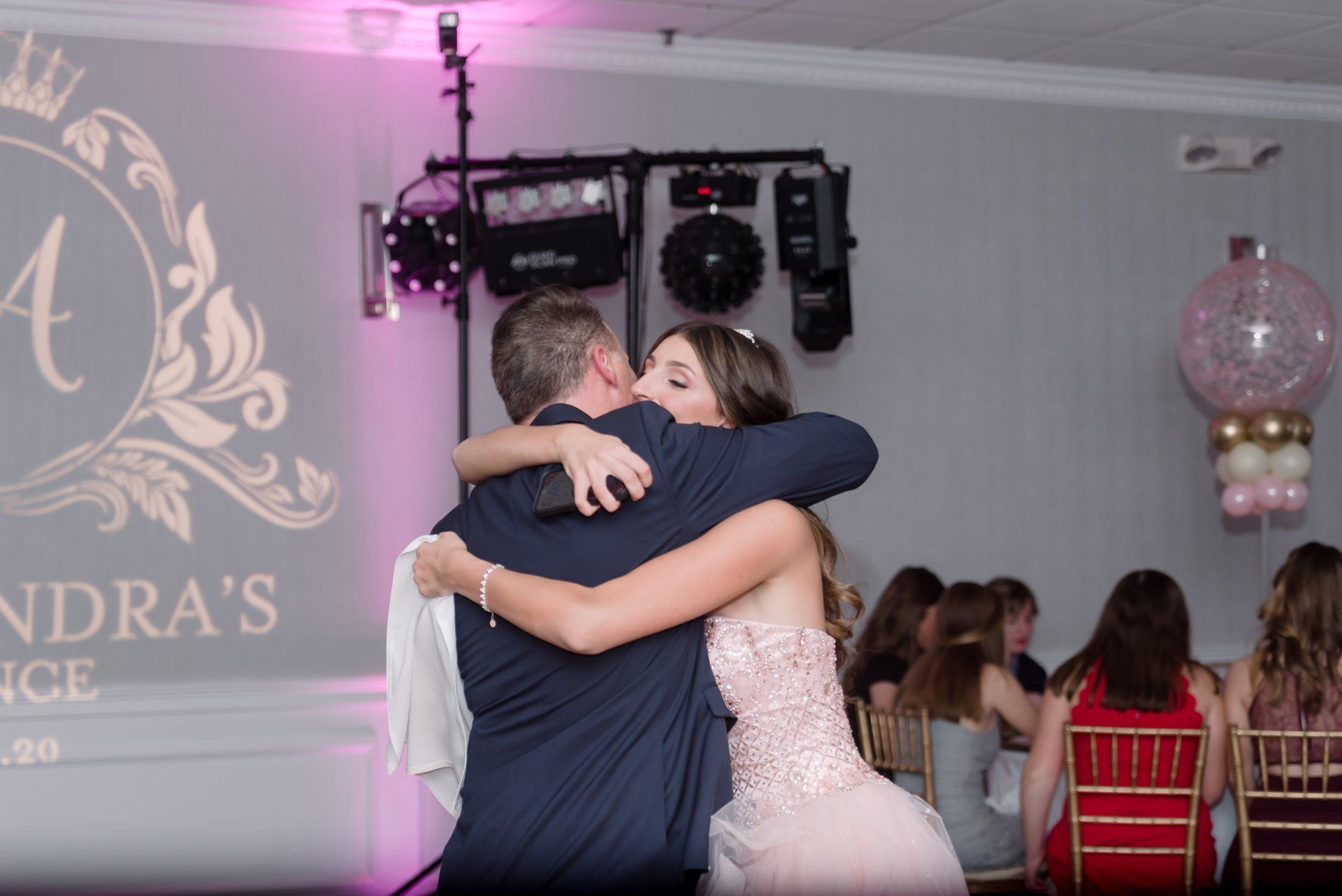 Two people hugging on a dance floor at a formal event, with pink lighting and guests in the background.