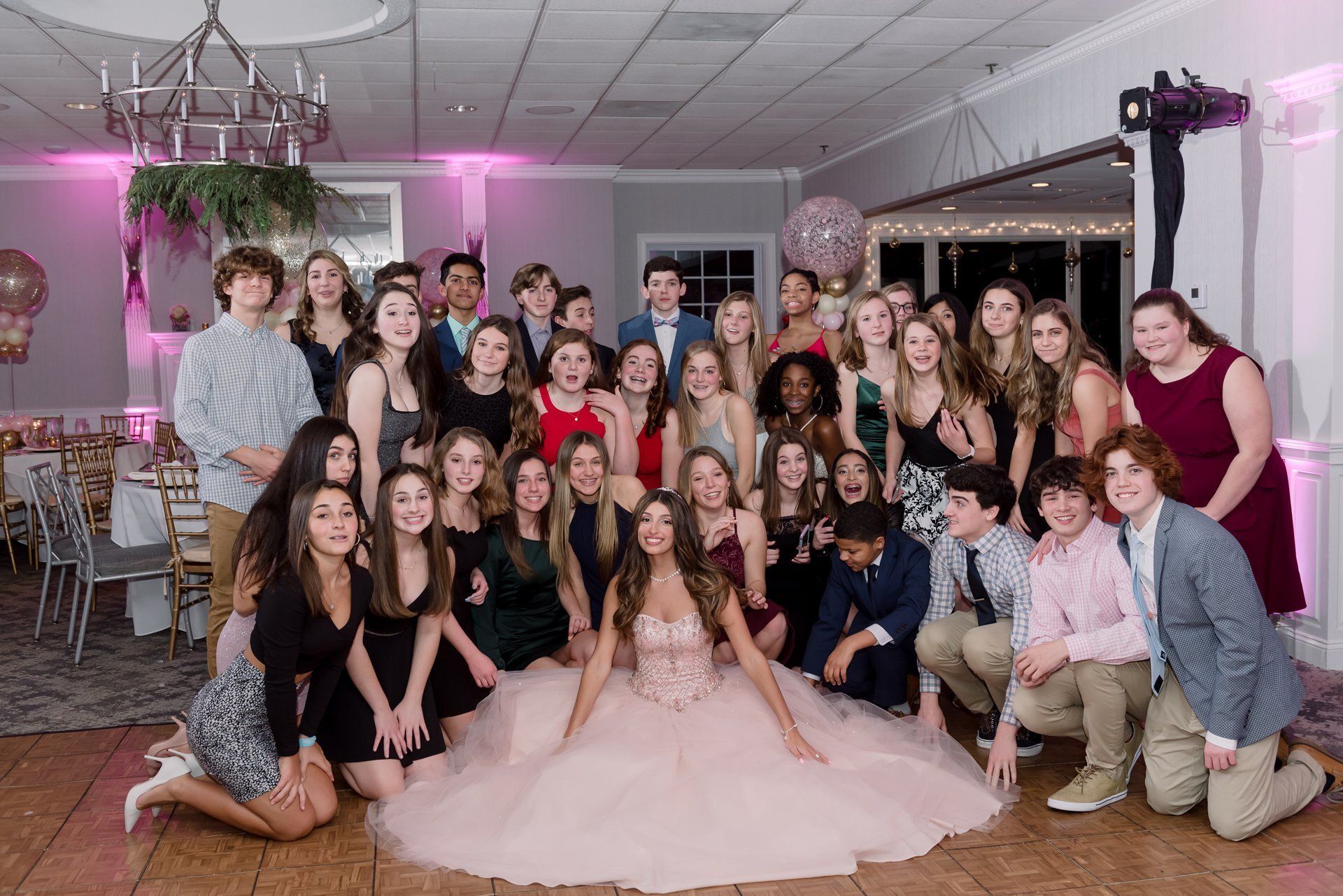 Large group posing in a decorated banquet hall, with a woman in a pink gown seated front center.
