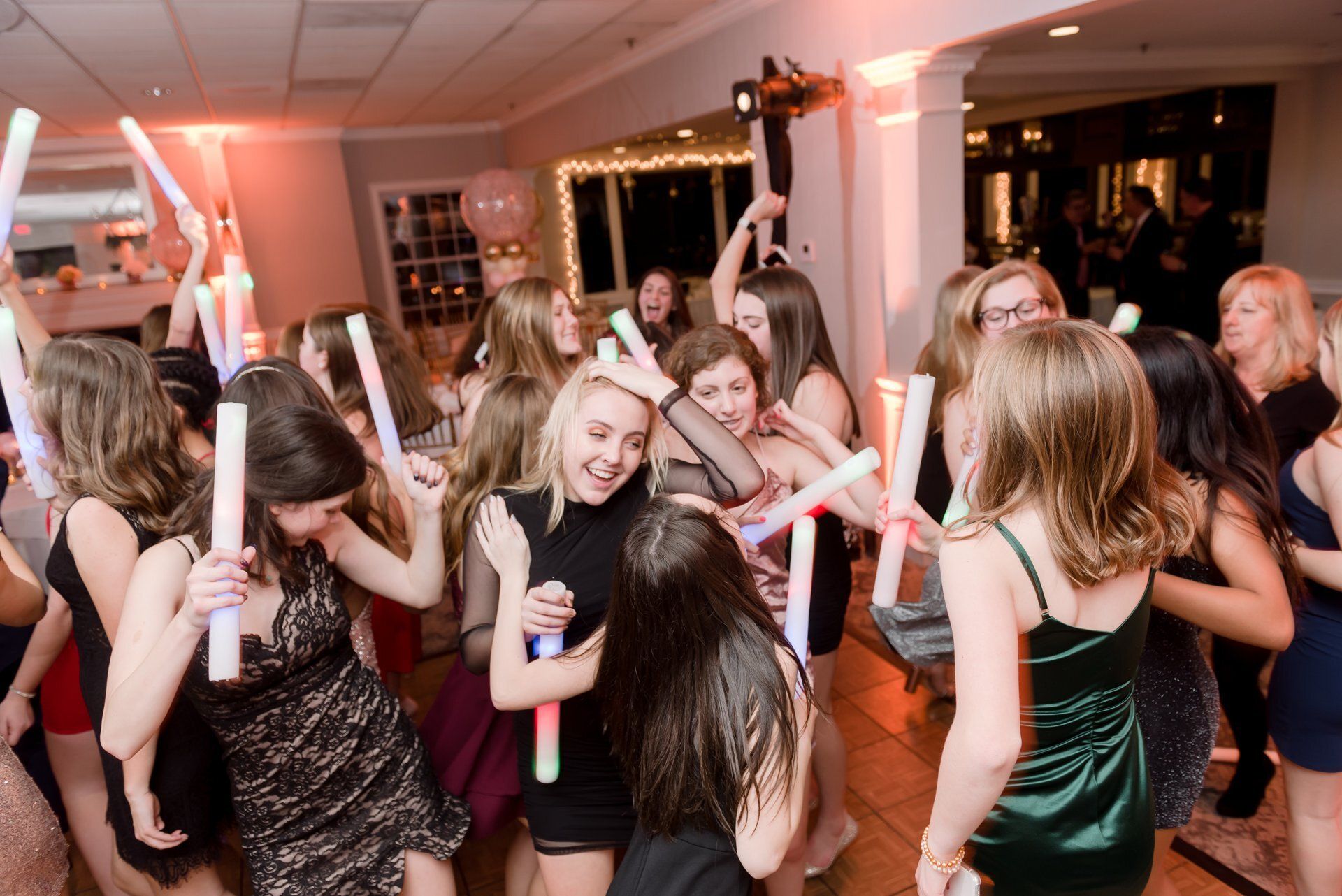 People dancing at a lively indoor party with glowing light sticks and warm string lights