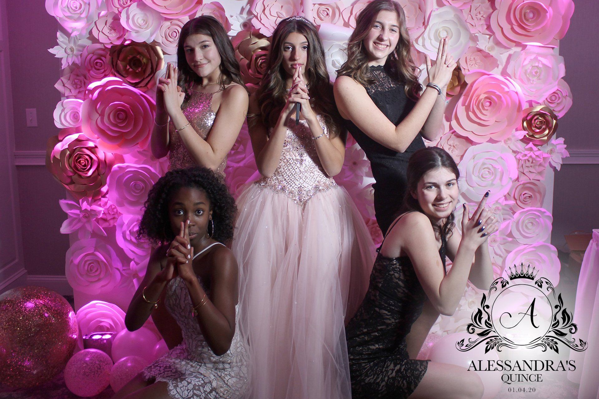 Five women pose in pink gowns against a floral rose backdrop, some holding flowers.