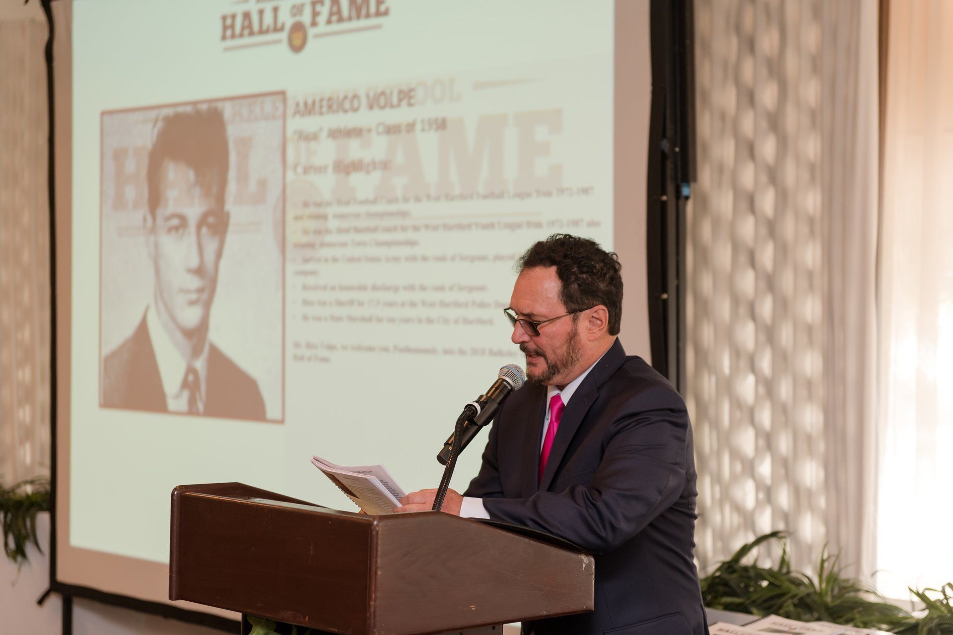 Man speaking at a podium during a hall of fame presentation, with a projected portrait and text behind him