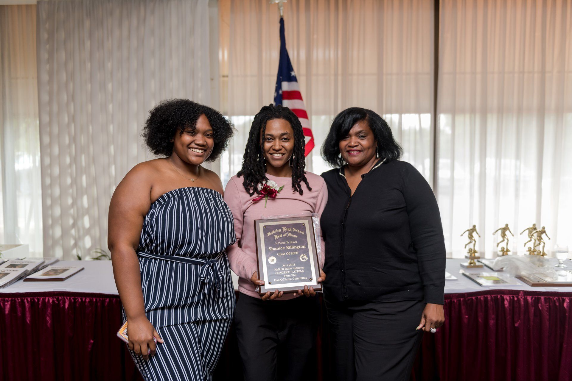 Three people posing indoors with an award plaque in front of curtains and an American flag.