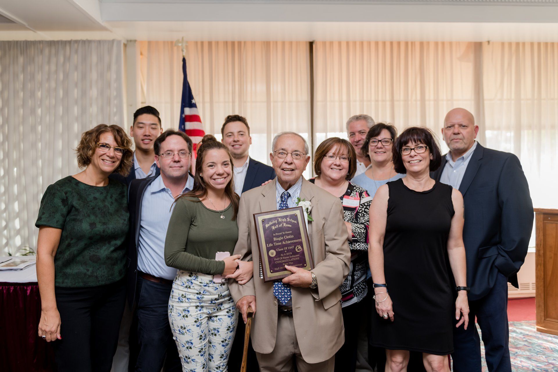 Group posing indoors with a framed plaque, standing in front of a U.S. flag and curtains