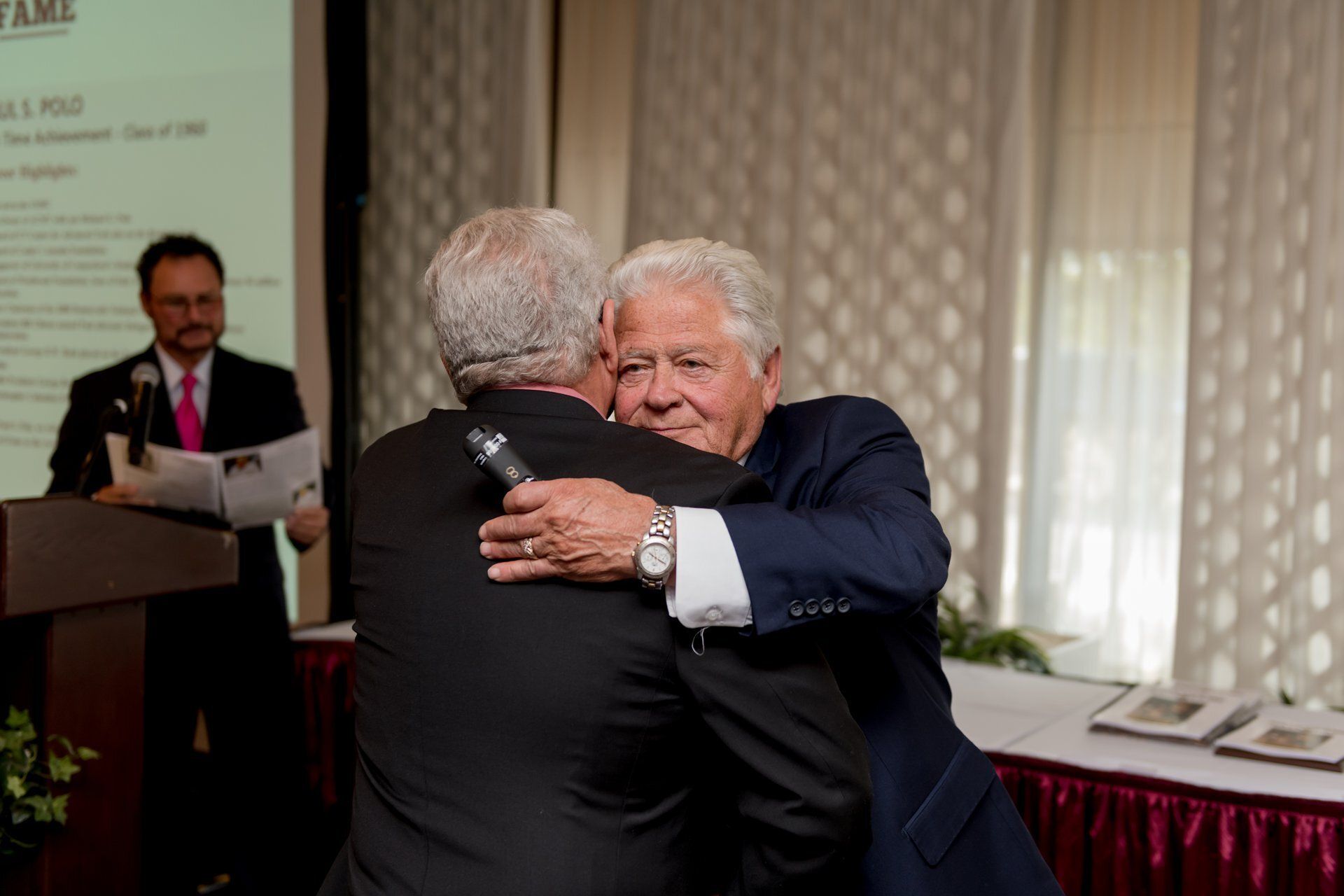Two men in suits embrace at a formal indoor event, with a speaker at a podium in the background.