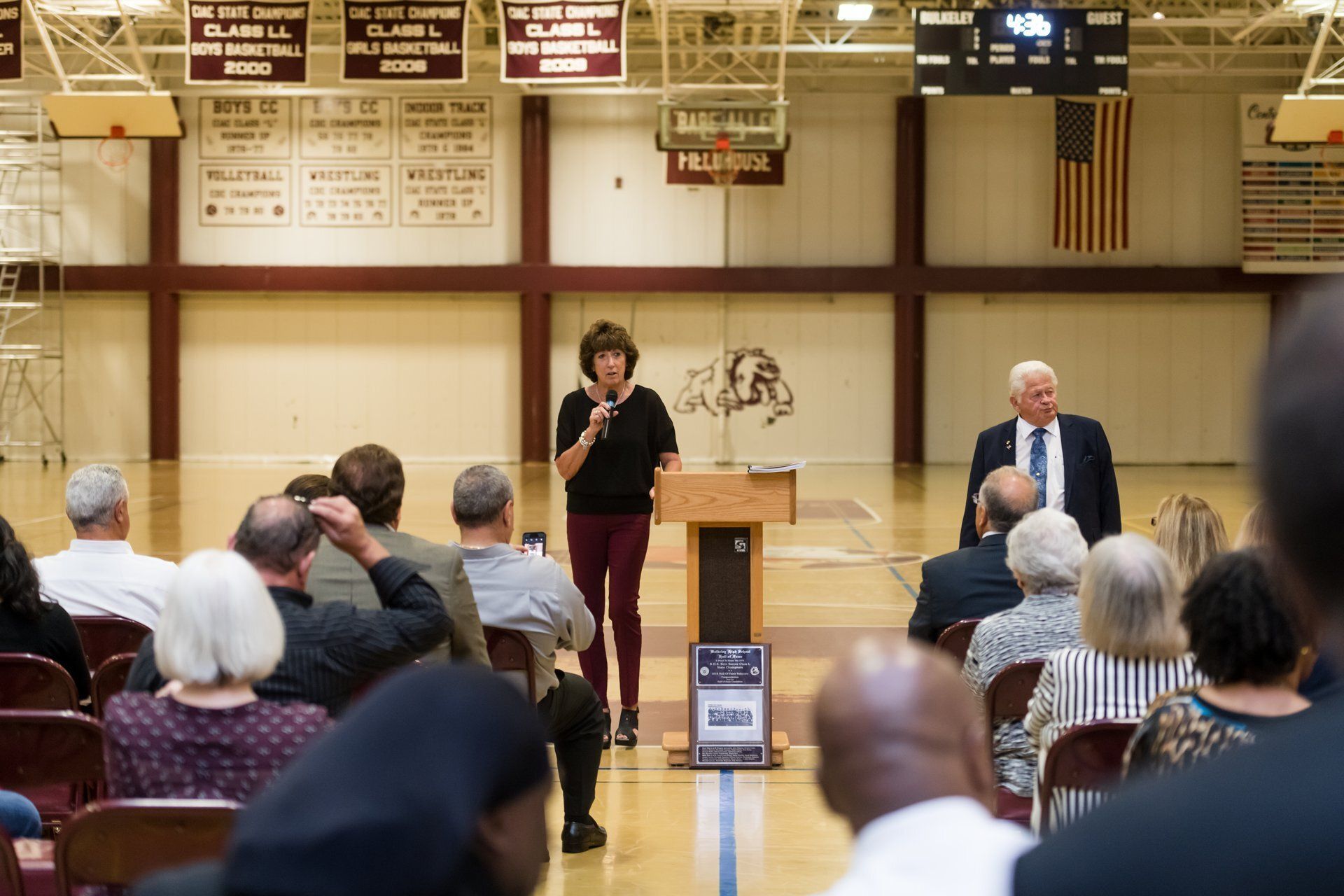 Speaker at podium addresses seated audience in a gymnasium with banners and an American flag.