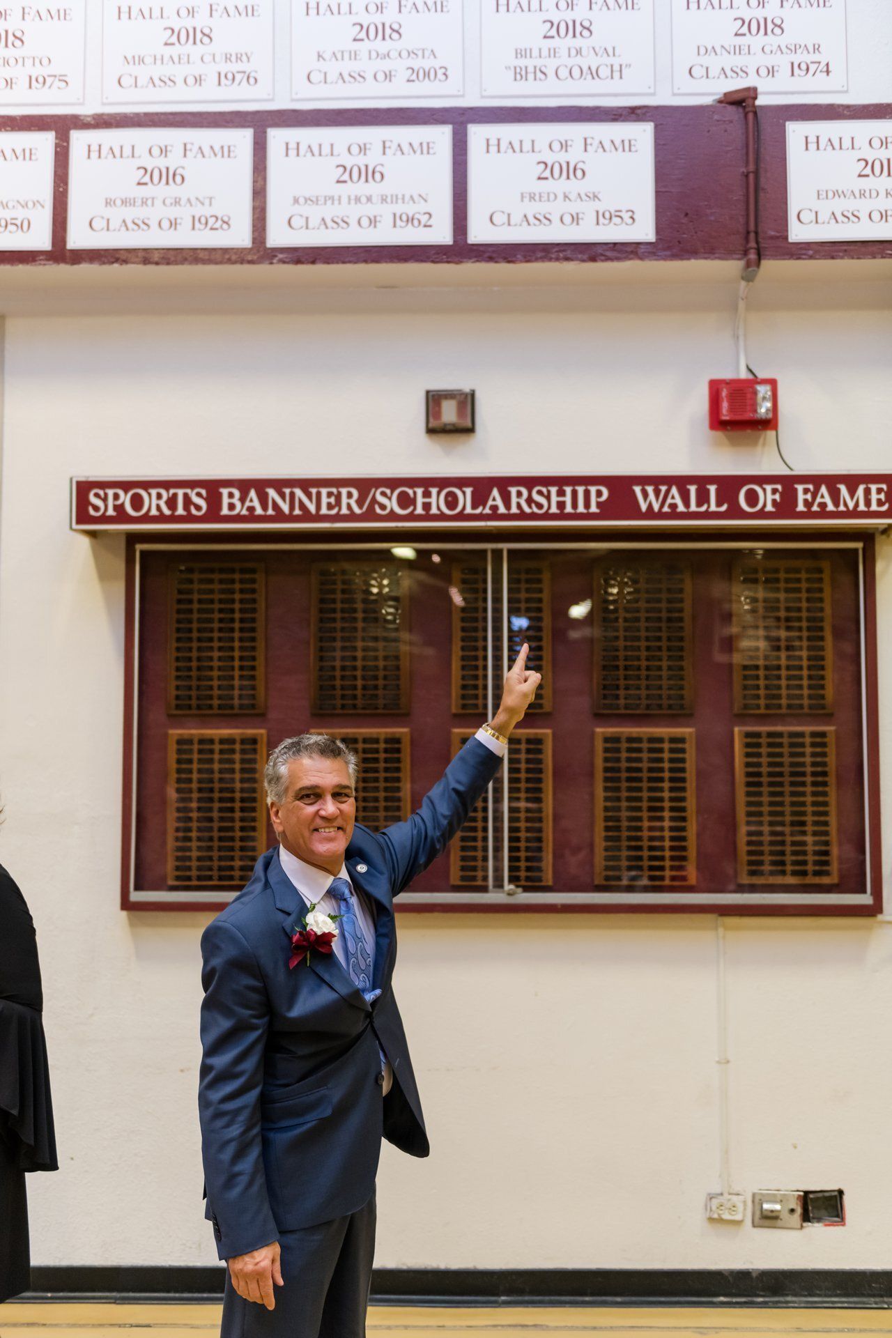 Man in a blue suit pointing up at a wall display labeled “Sports Banner Scholarship Hall of Fame”