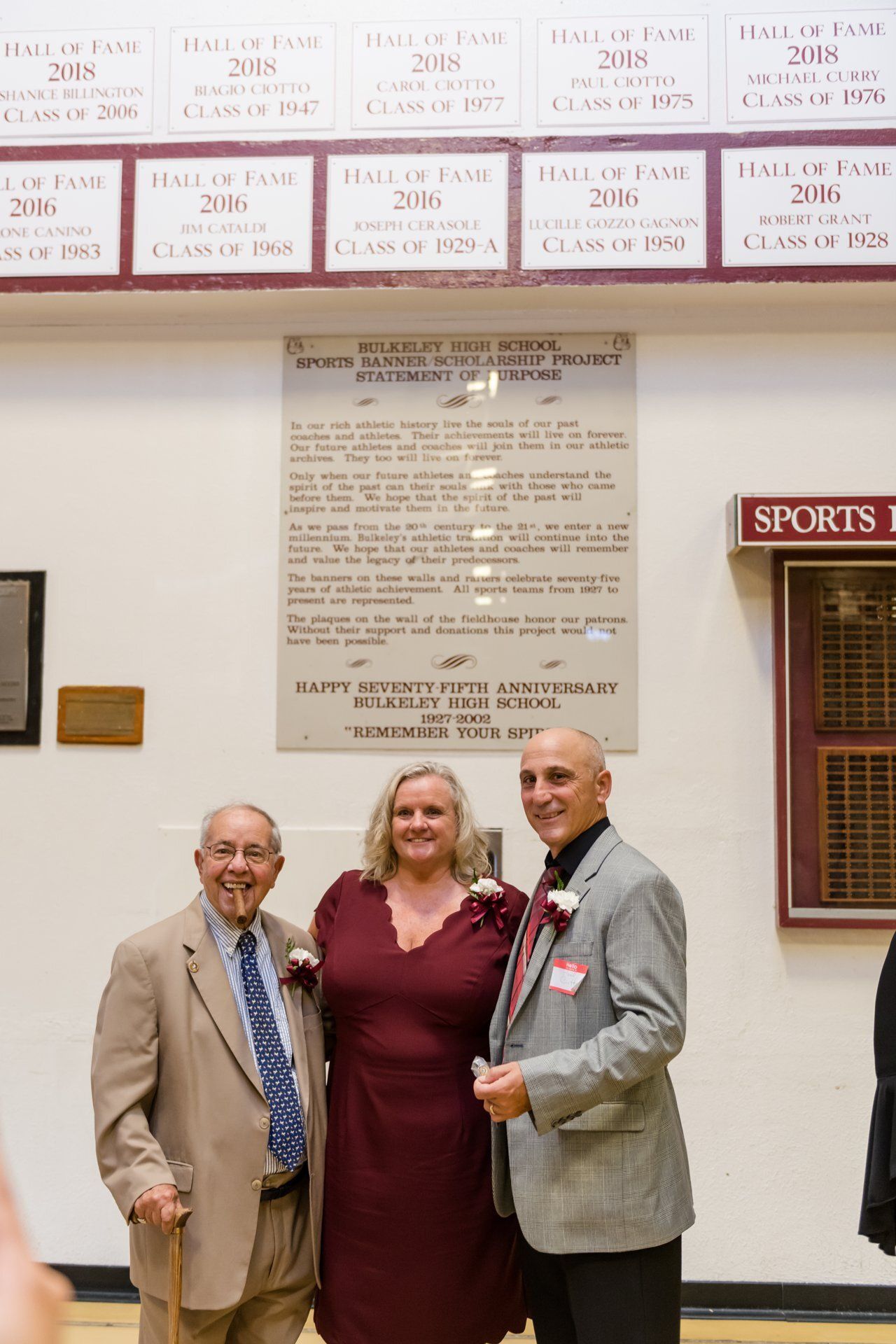 Three people posing indoors beside a commemorative plaque and sports display.