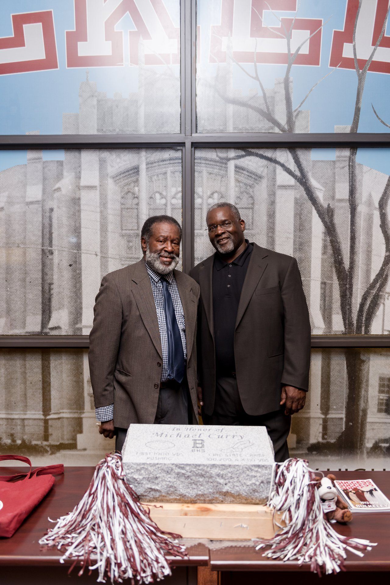 Two men standing behind a table with a cake and red-and-white pom-poms in front of a glass wall