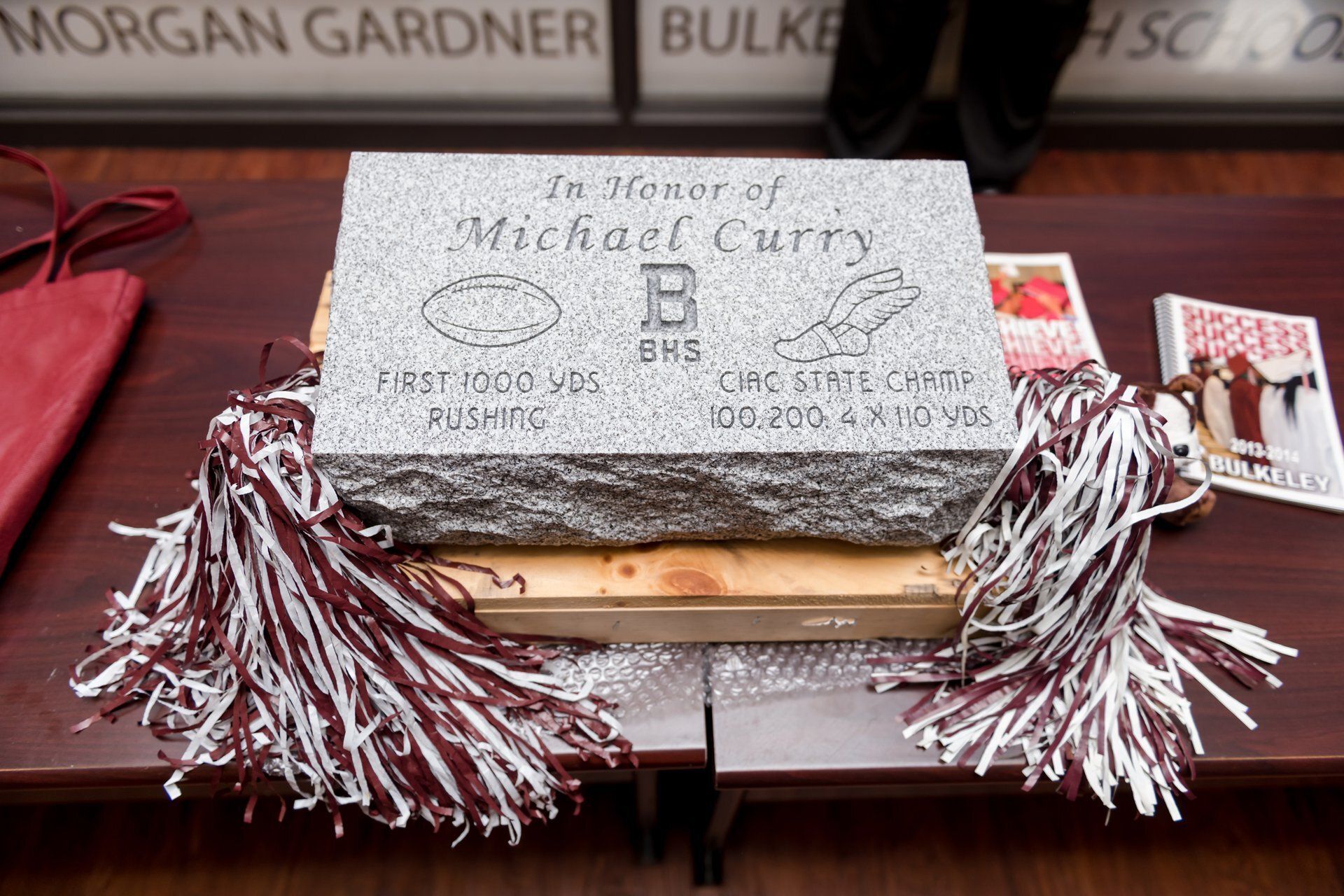 White graduate cap with maroon-and-white tassels and “Michael Curry” written on top, displayed on a table