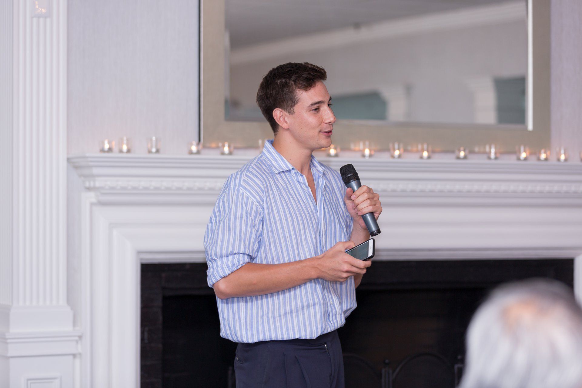 Man speaking into a microphone in a bright room with a fireplace and candles behind him
