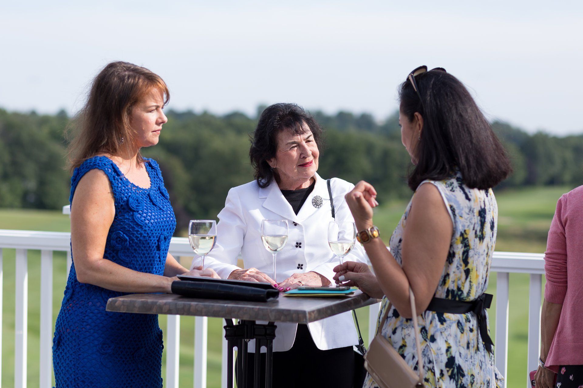 Three women converse at a cocktail table outdoors near a railing, holding drinks in a green field.