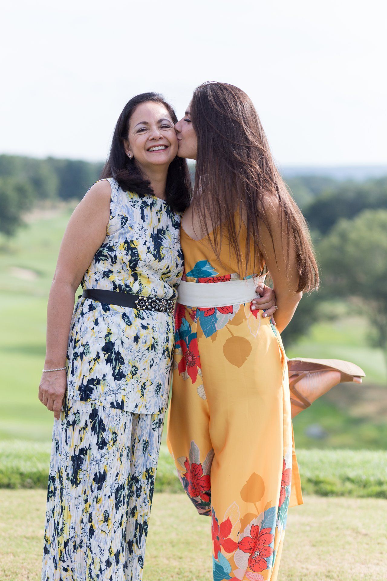 Two women smiling and embracing outdoors in colorful dresses, with a grassy landscape behind them.