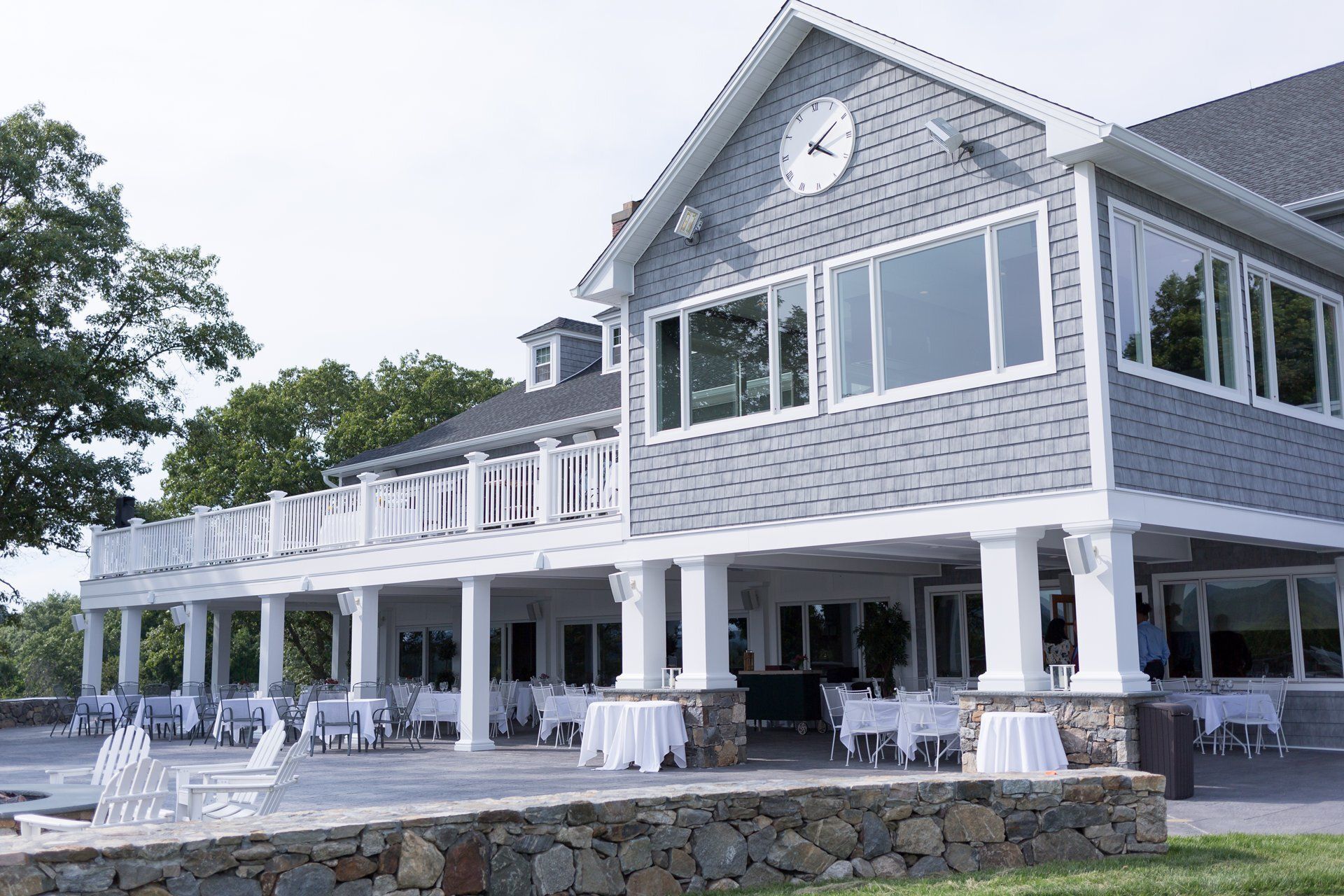Large gray shingle building with white trim, clock on gable, and outdoor seating under a covered porch by stone wall