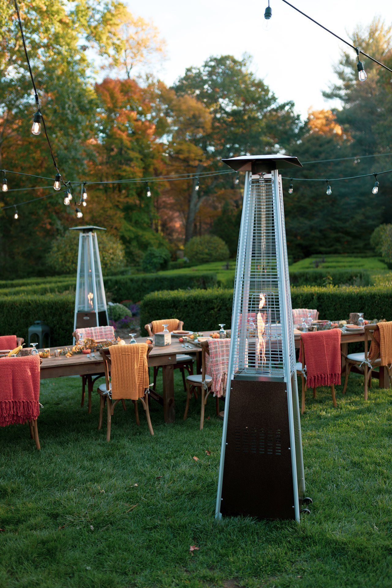 Outdoor patio dining setup with tall patio heaters, string lights, and autumn trees in the background