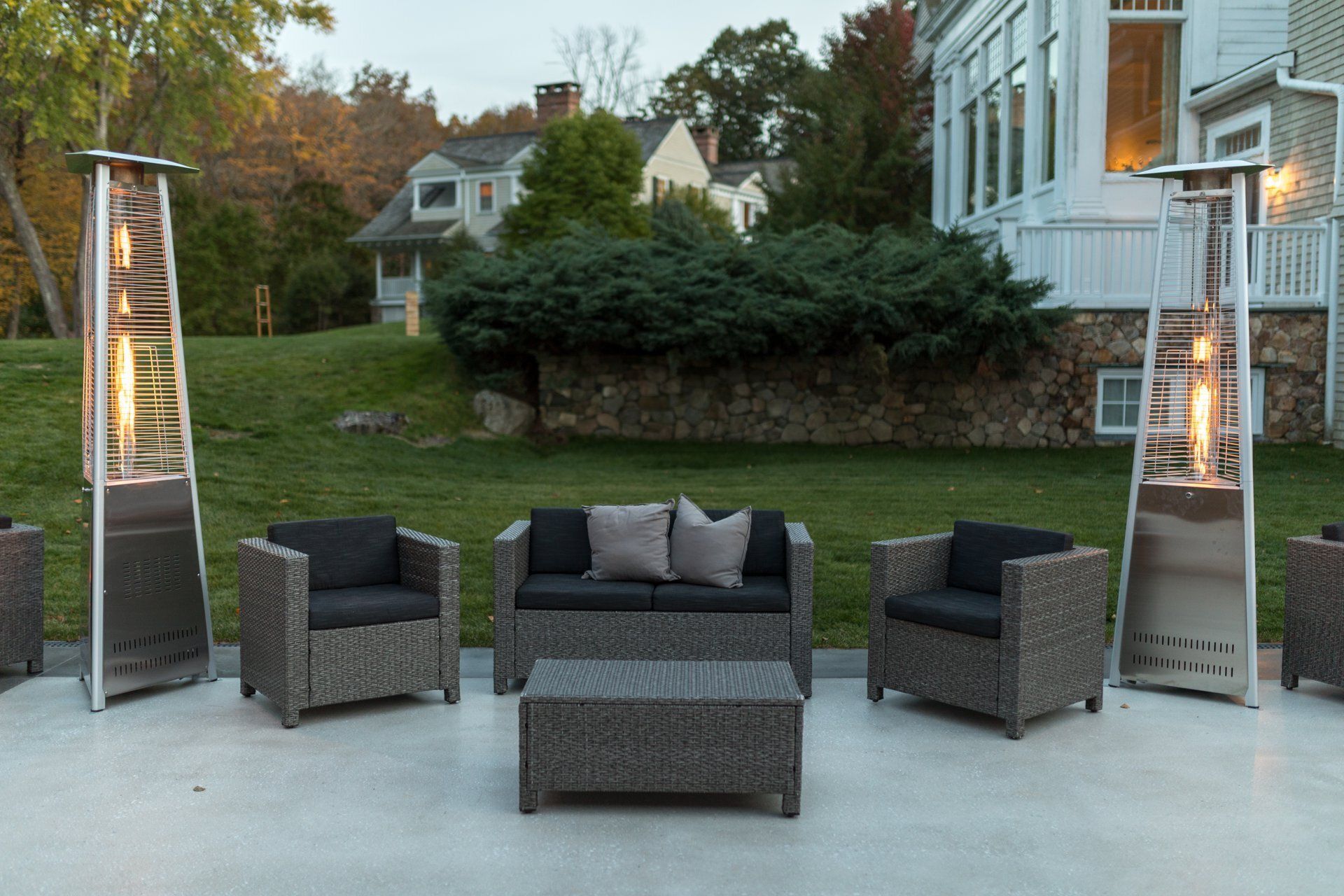Outdoor patio with four dark wicker chairs, a coffee table, and two tall patio heaters at dusk.