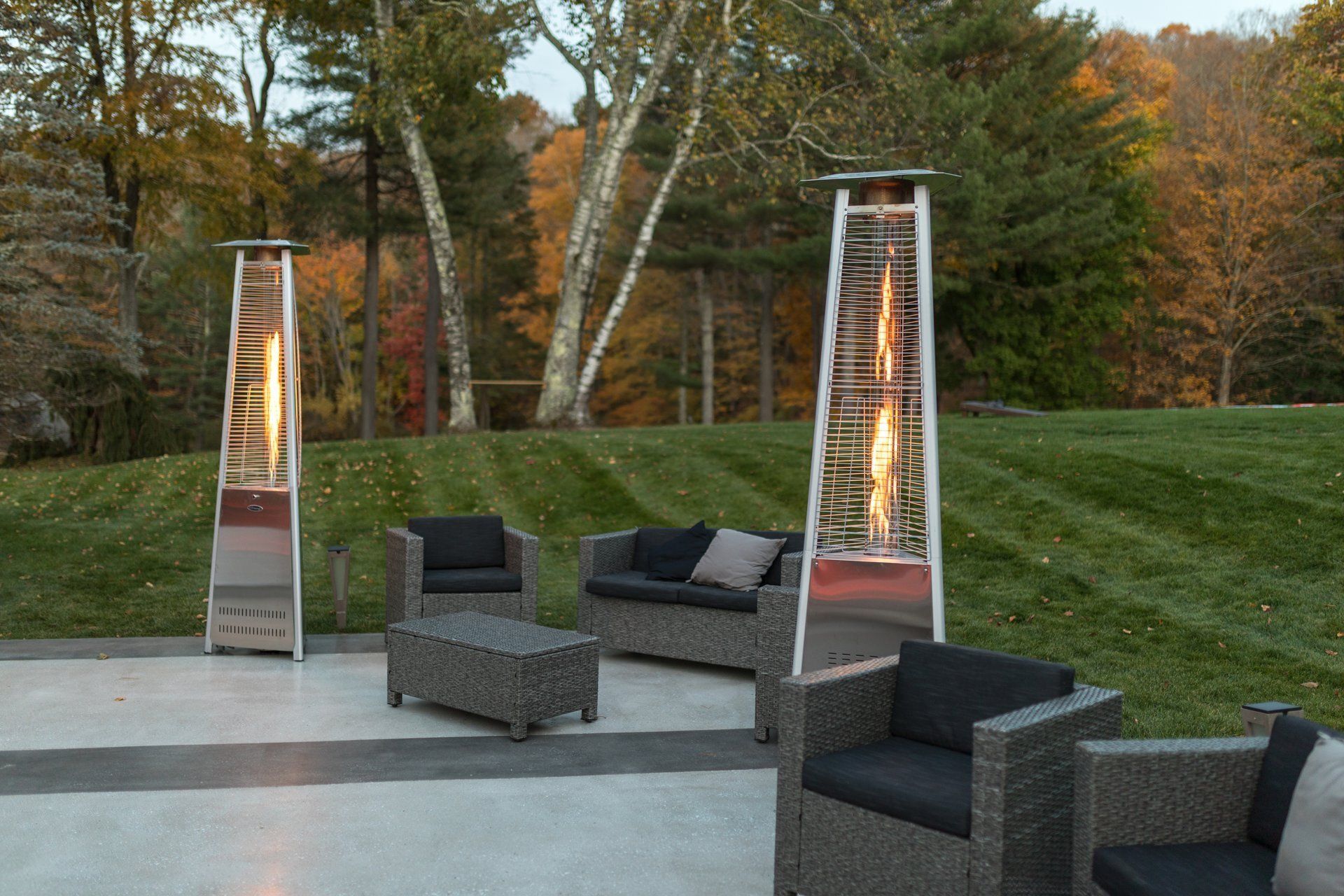 Outdoor patio with wicker lounge chairs and lit tower heaters beside a grassy hillside and autumn trees