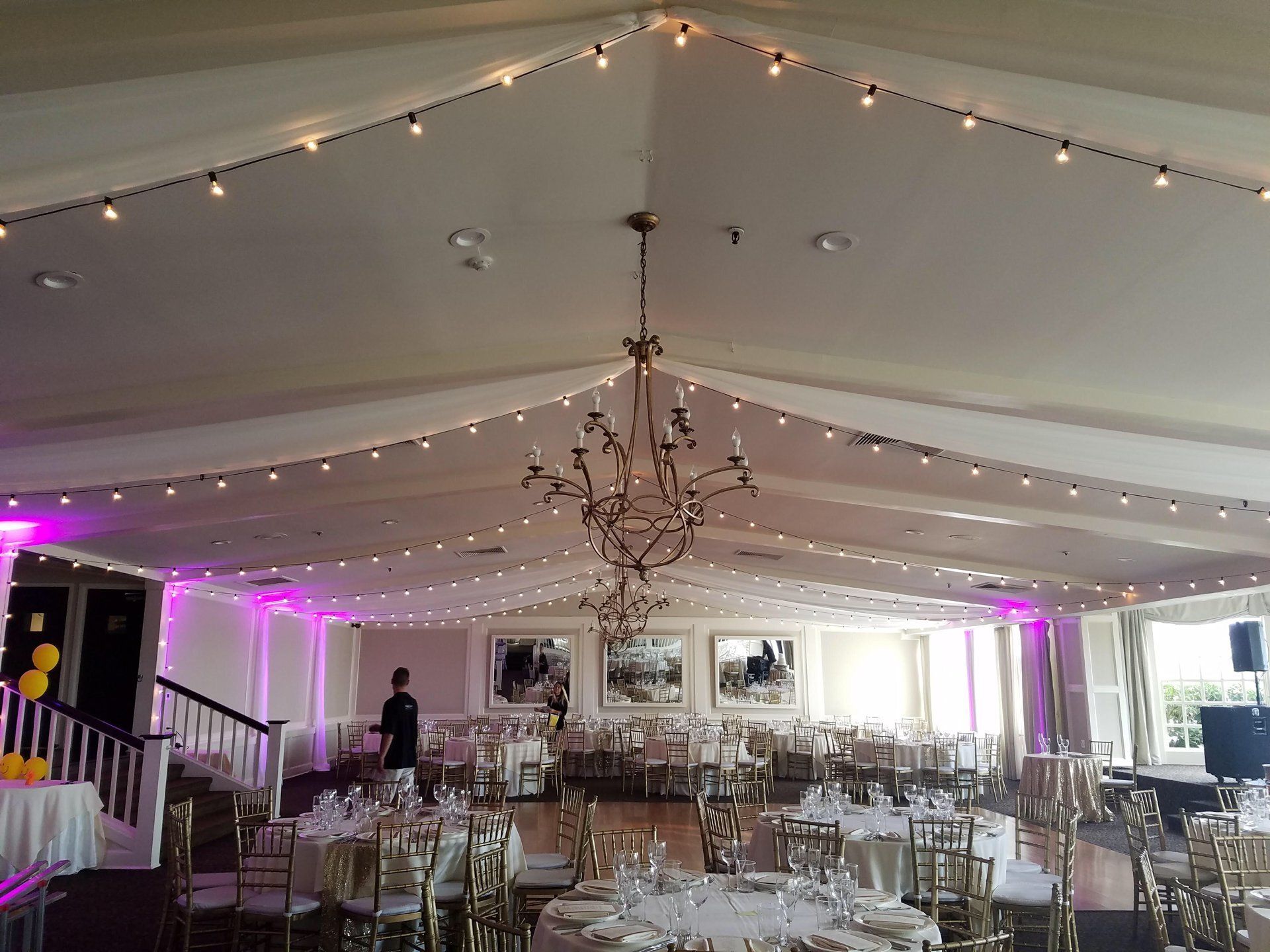 Elegant banquet hall with chandeliers, string lights, and rows of gold chairs and tables under a draped tent ceiling