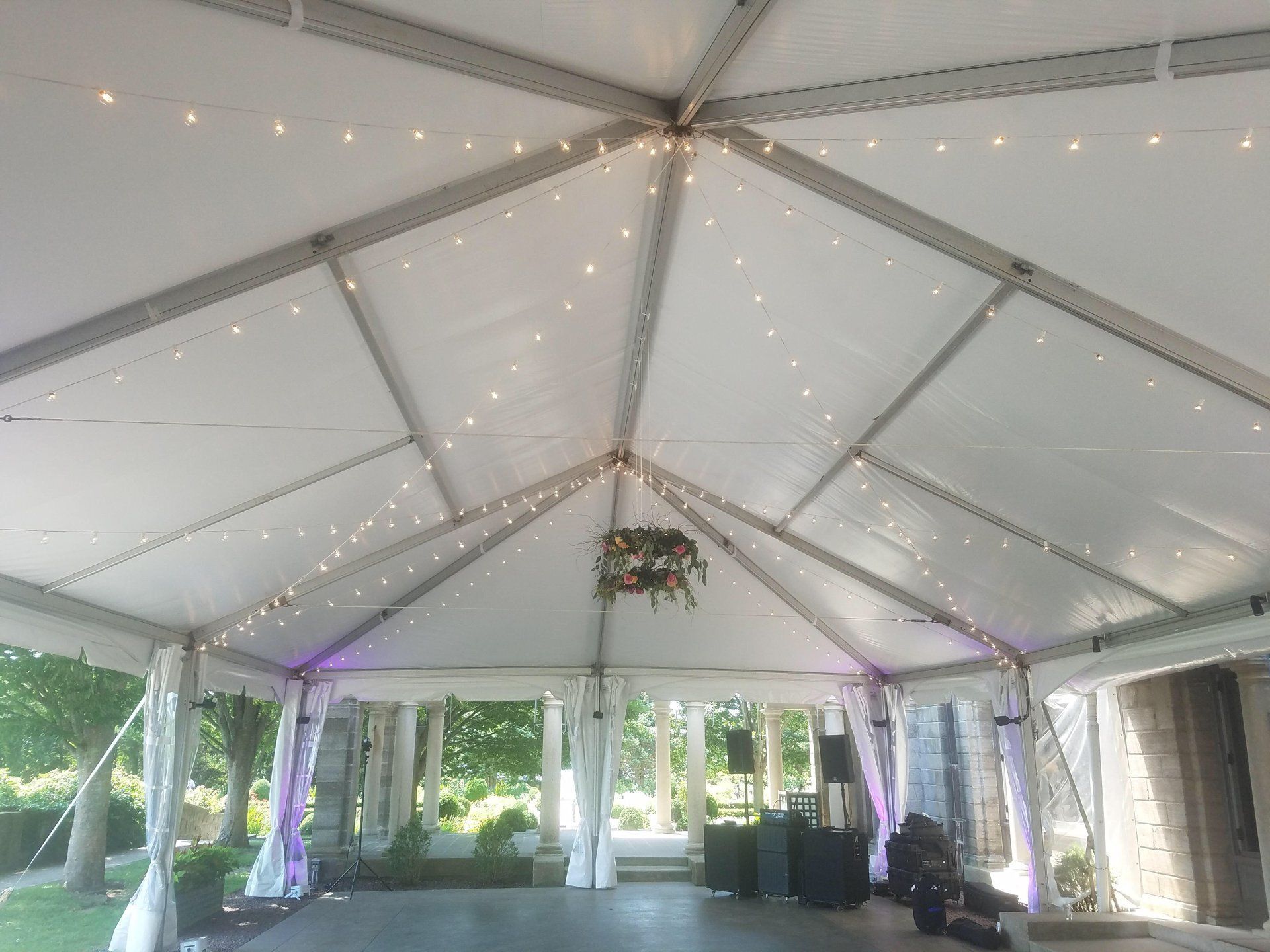 White event tent with string lights and hanging greenery, open sides overlooking a grassy outdoor setting.