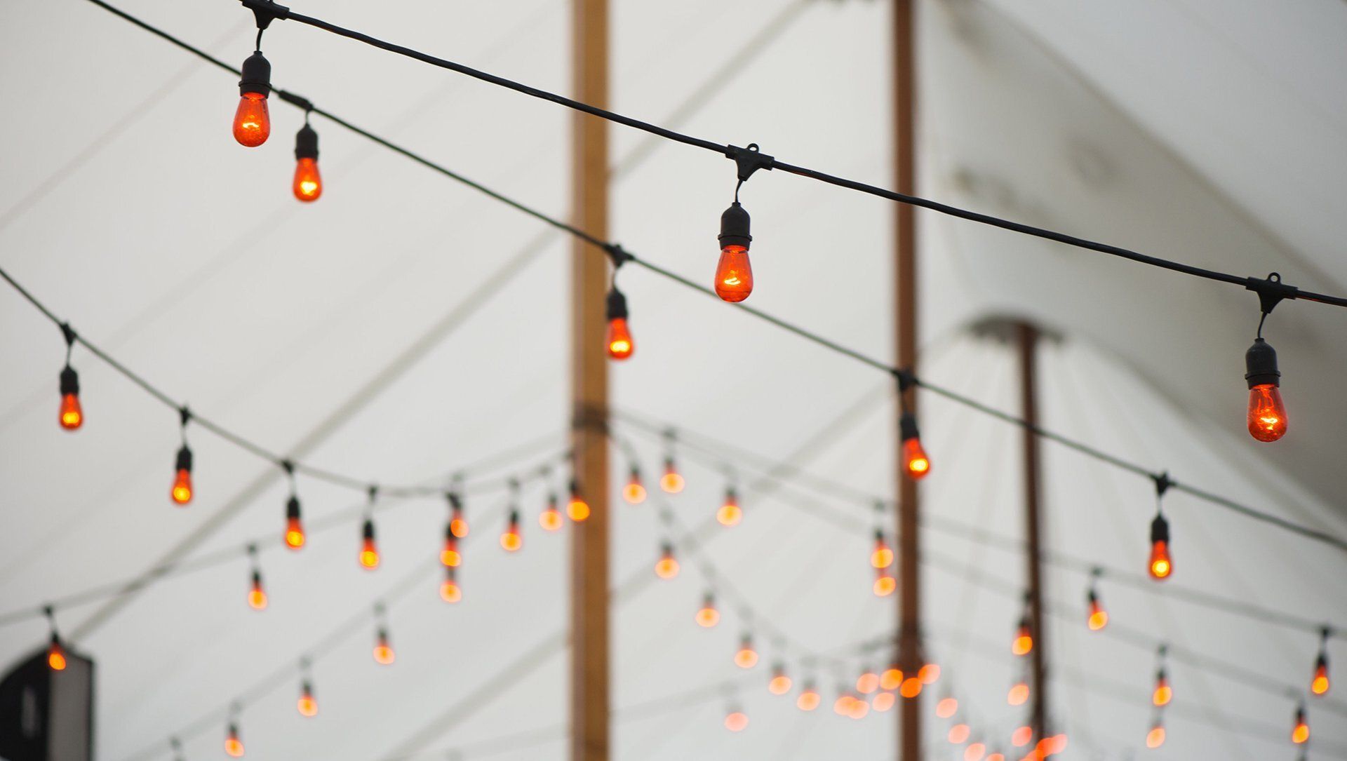 String lights with glowing orange bulbs hang across a white tented outdoor space.