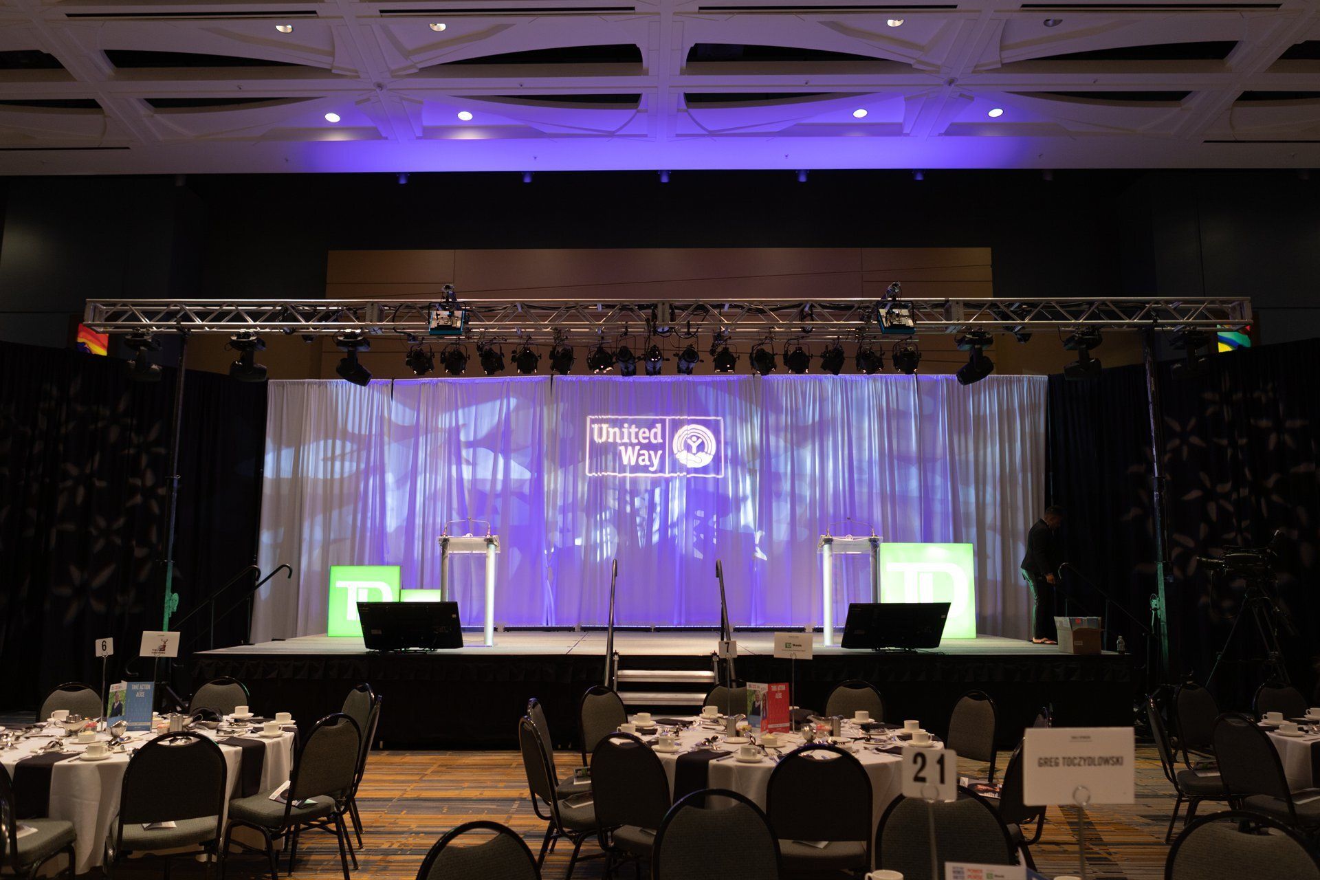 Empty banquet hall with a lit stage, purple backdrop, and tables set in the foreground
