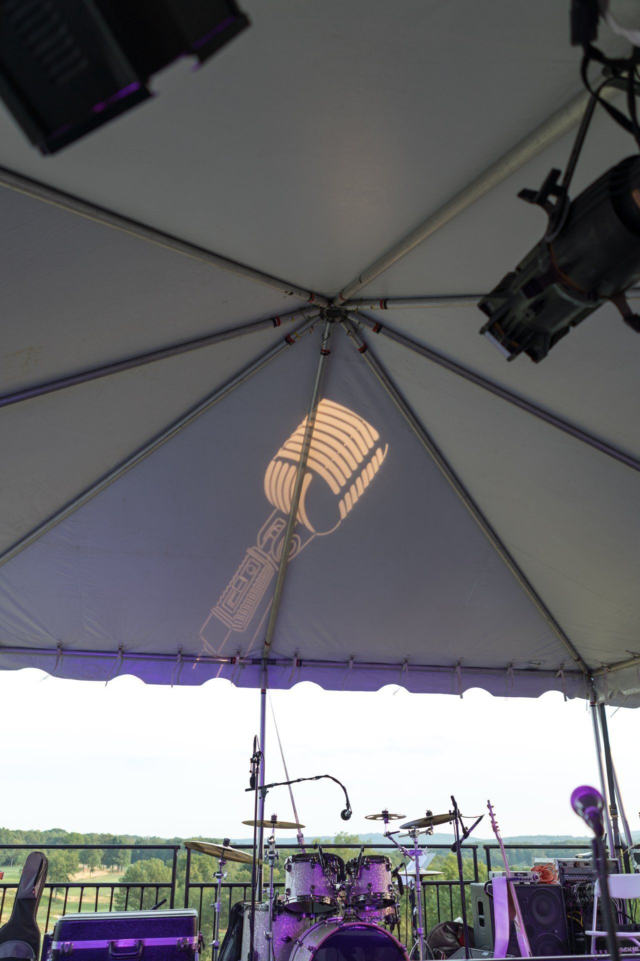 Under a white tent, a stage setup with purple flowers, microphones, and sunlight shining through the canopy.
