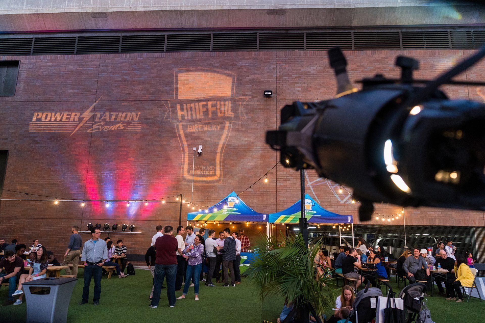 Outdoor evening festival crowd under colorful tents beside a brick wall with projected lights and a large sign.