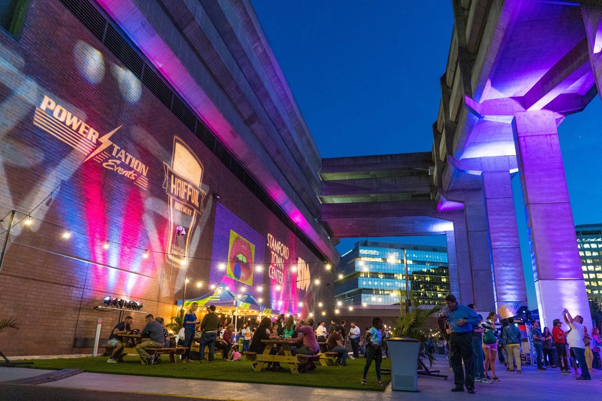 Nighttime outdoor event between modern buildings, lit with purple and pink lights, with a crowd gathered below.