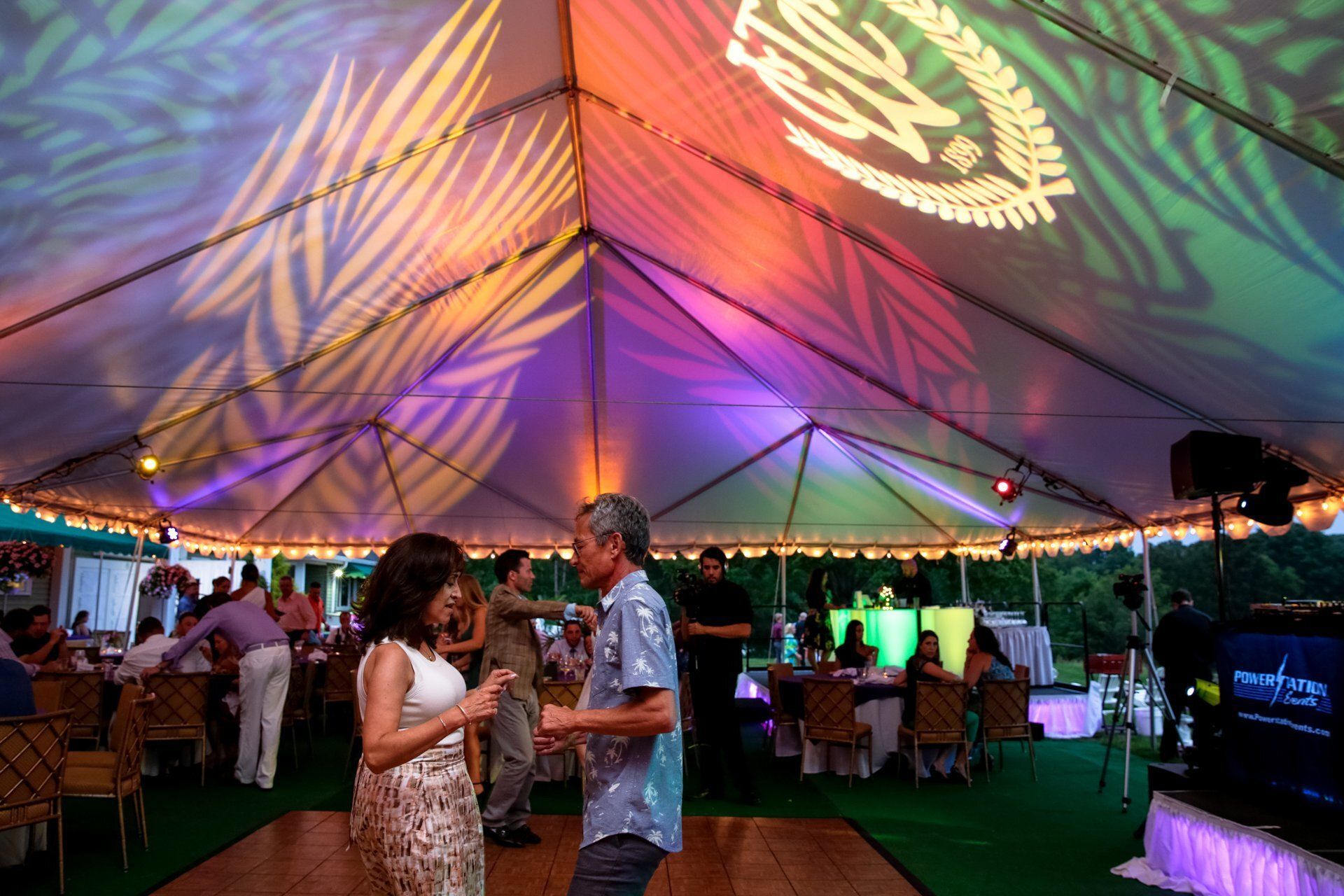 Couple dancing under a colorful tent at an outdoor reception with guests and tables nearby