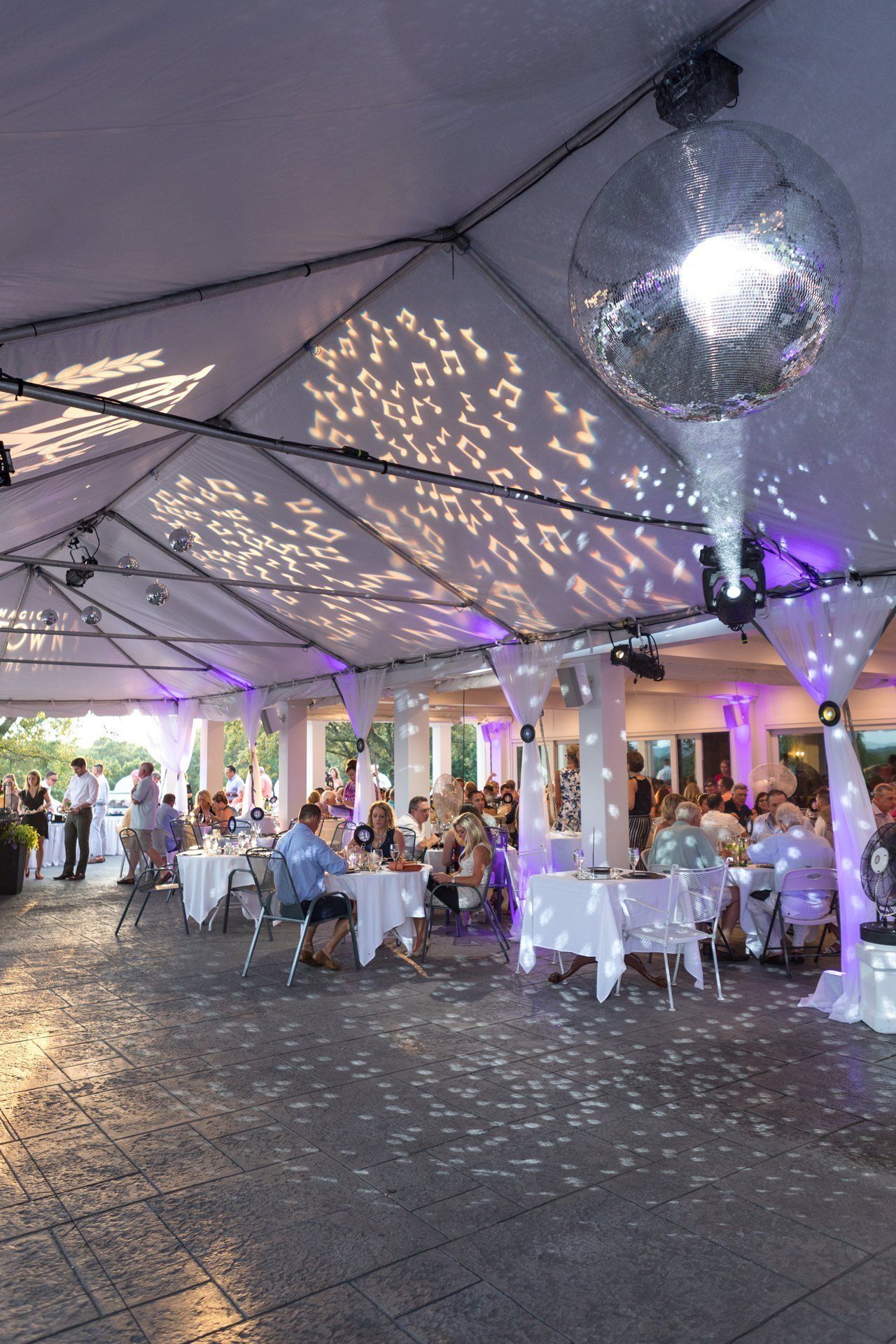 Elegant outdoor event under a white tent with seated guests, round tables, and disco ball lighting.