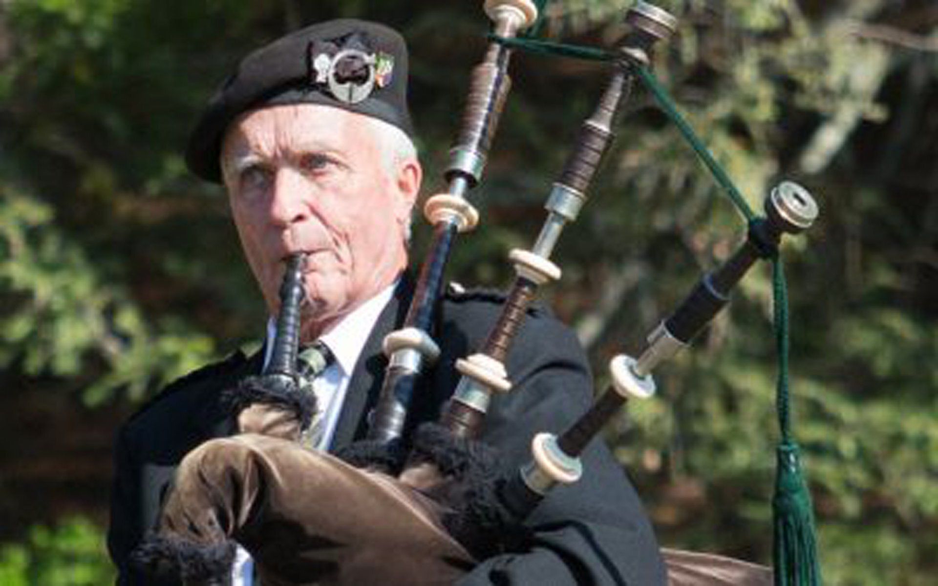 Man playing bagpipes outdoors in a black beret and dark jacket, with green trees in the background