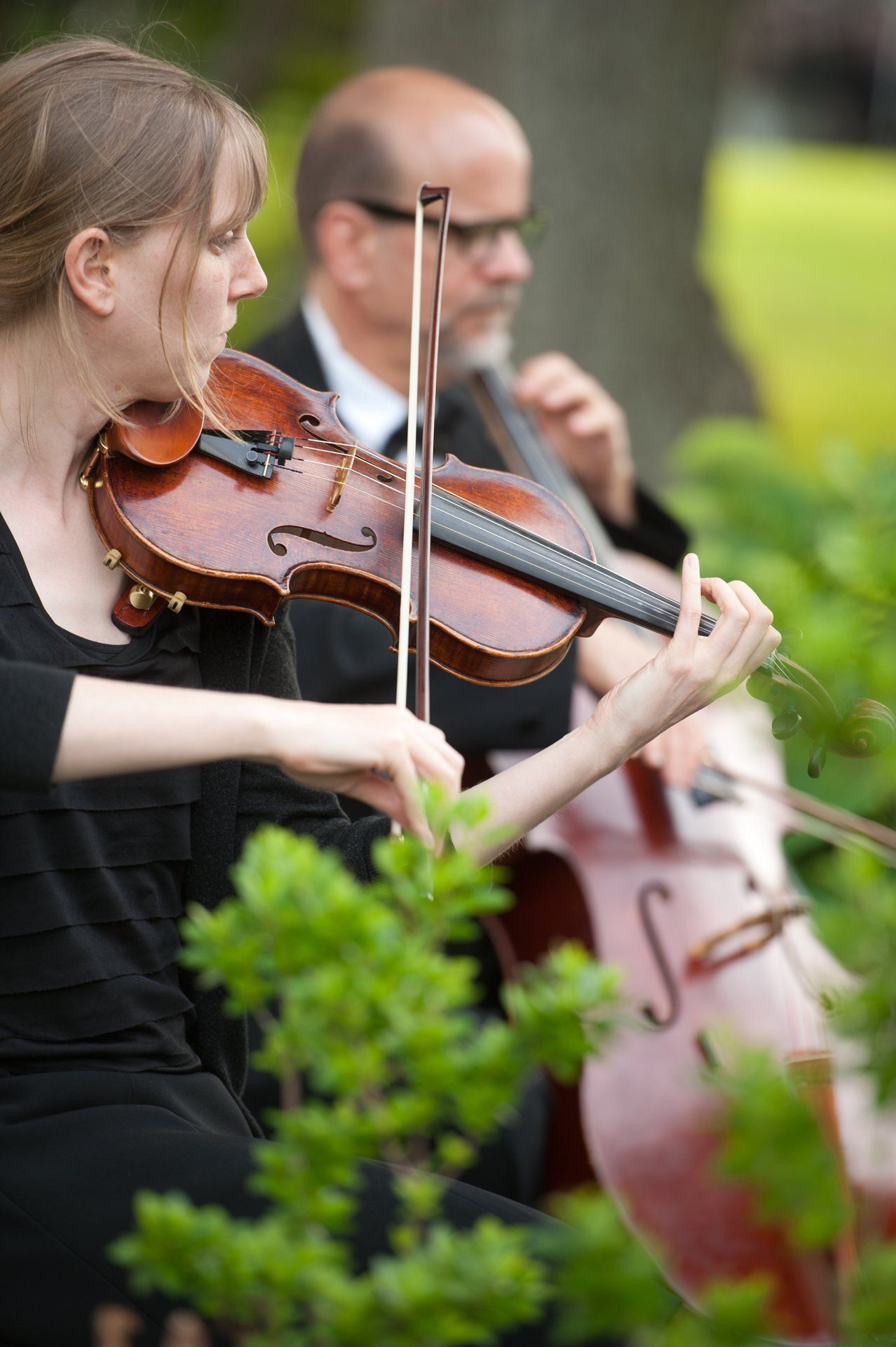 Two violinists performing outdoors, one in black clothing in the foreground and one blurred in the background.