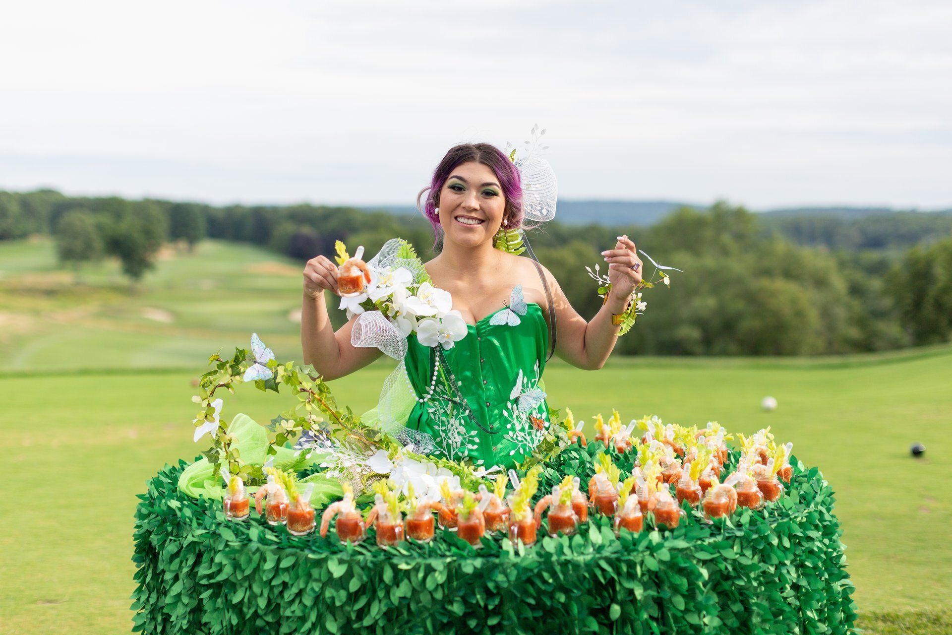 Woman in a green dress smiling behind a flower-covered table outdoors on a sunny field.
