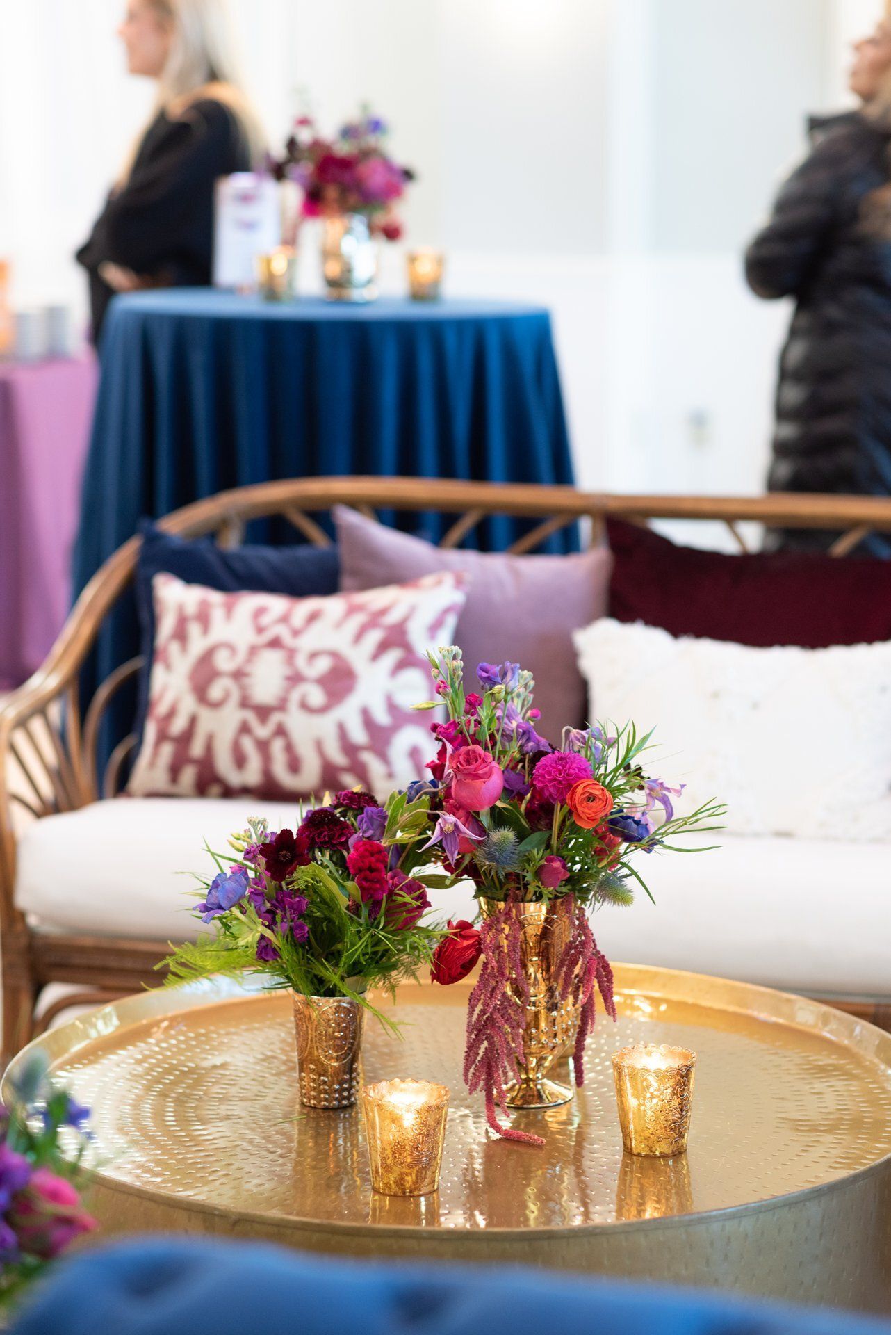 Decorative lounge with wicker sofa, pink cushions, floral arrangement, and gold tray table at an event