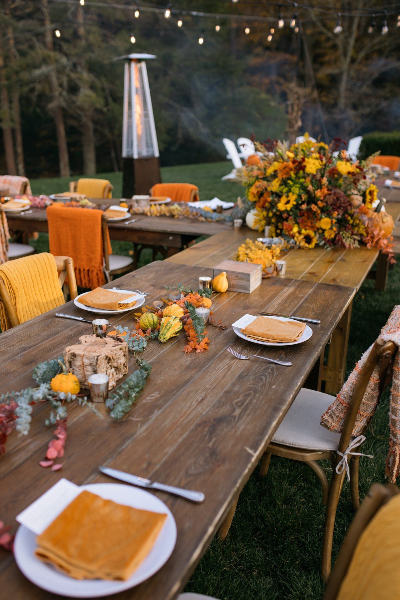 Rustic outdoor dining table set with orange accents, candles, and autumn floral centerpiece under string lights