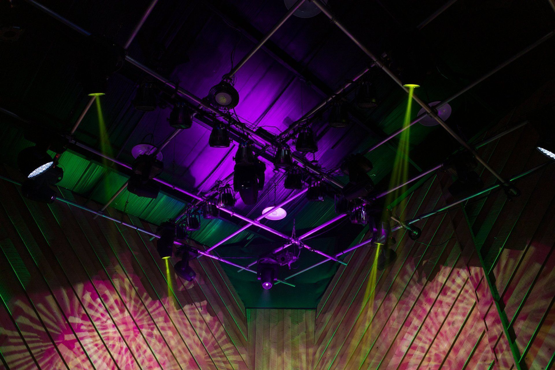 Purple and green laser lights over a concert stage with hanging rigging and beams across the ceiling