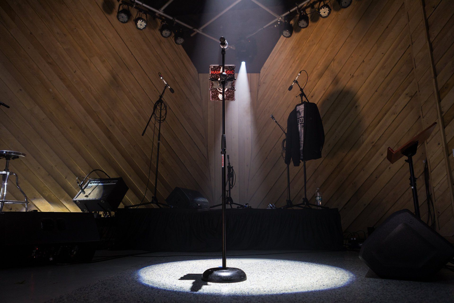 Empty stage with a central microphone stand under a spotlight, flanked by a coat rack and speakers.