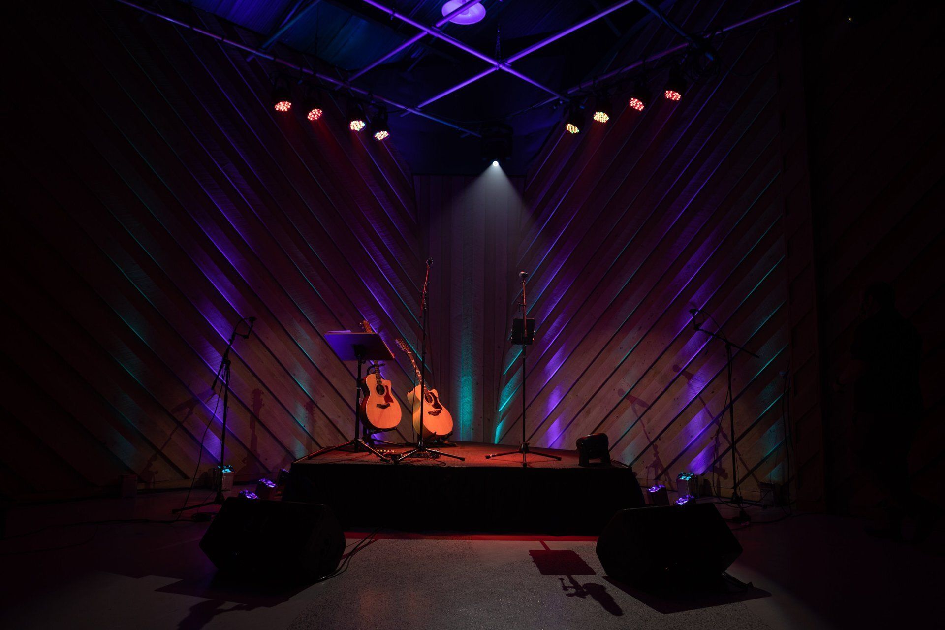 Dimly lit stage with two guitars on stands, colorful spotlights, and a microphone in the center