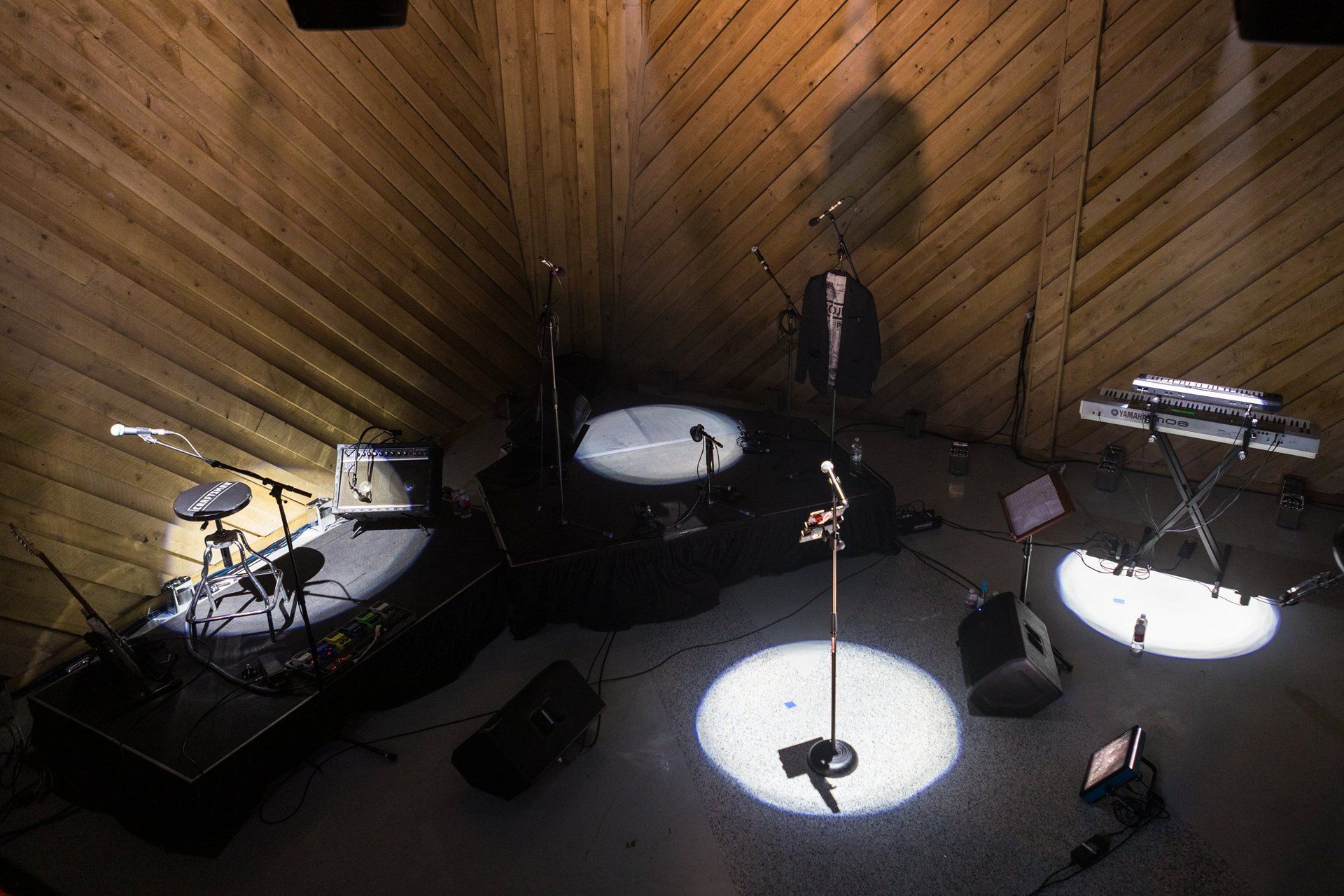 Dim rehearsal room with drum kits, cymbals, microphones, and a wooden slanted ceiling.
