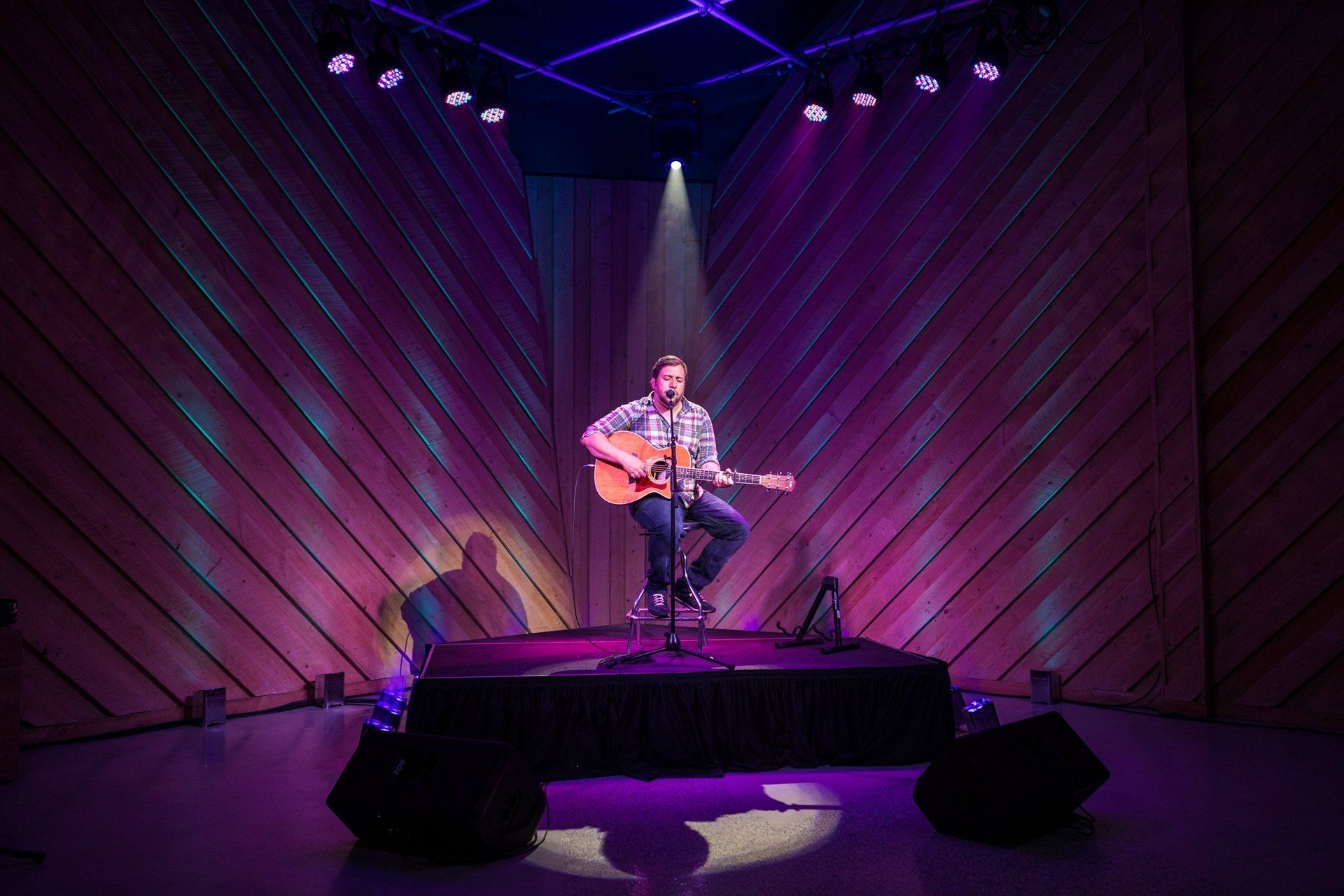Musician seated on a lit stage, playing acoustic guitar under purple spotlights.