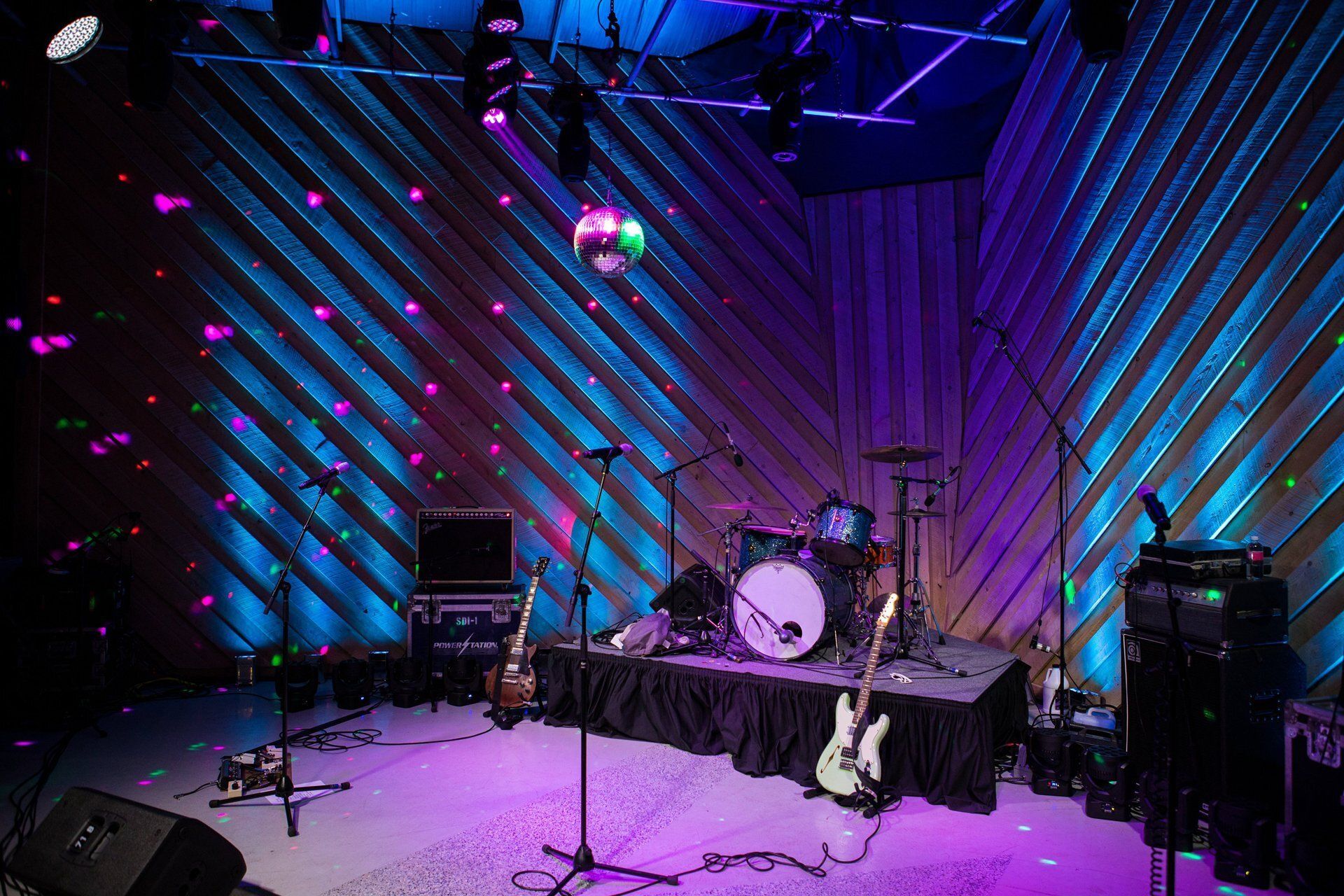 Empty stage with drum kit and guitars under purple-blue lighting in a small venue
